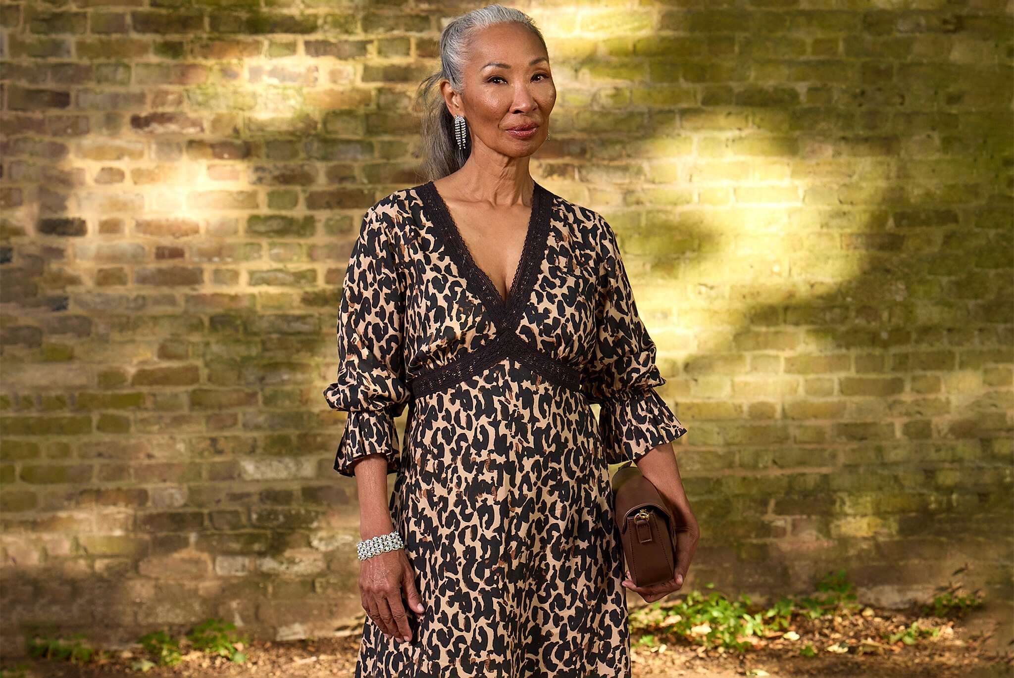 Person wearing a leopard-print V-neck midi dress, holding a brown clutch, posed outdoors against a brick wall.