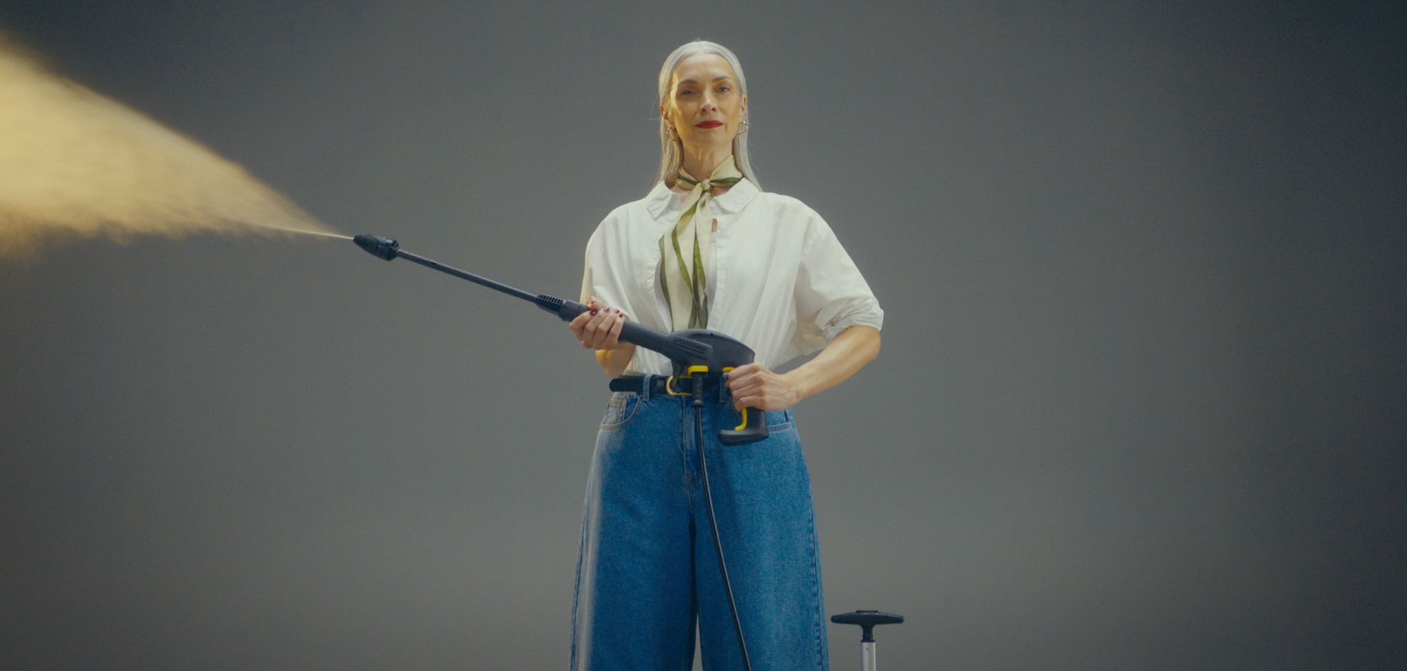 A woman wearing a white top and blue jeans holding a jet washer lance spraying water.