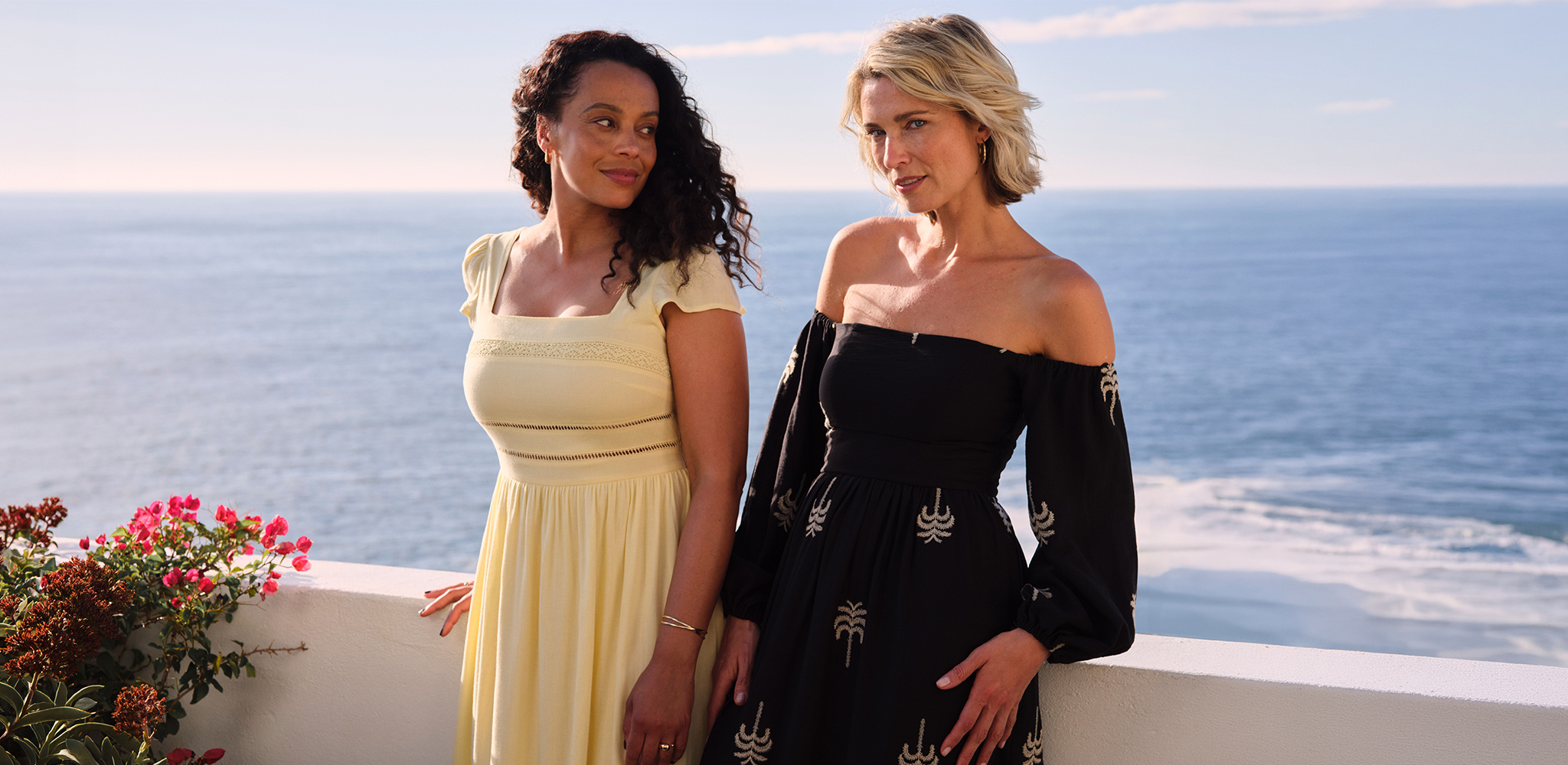 2 women wearing dresses stood next to a balcony at the sea side.