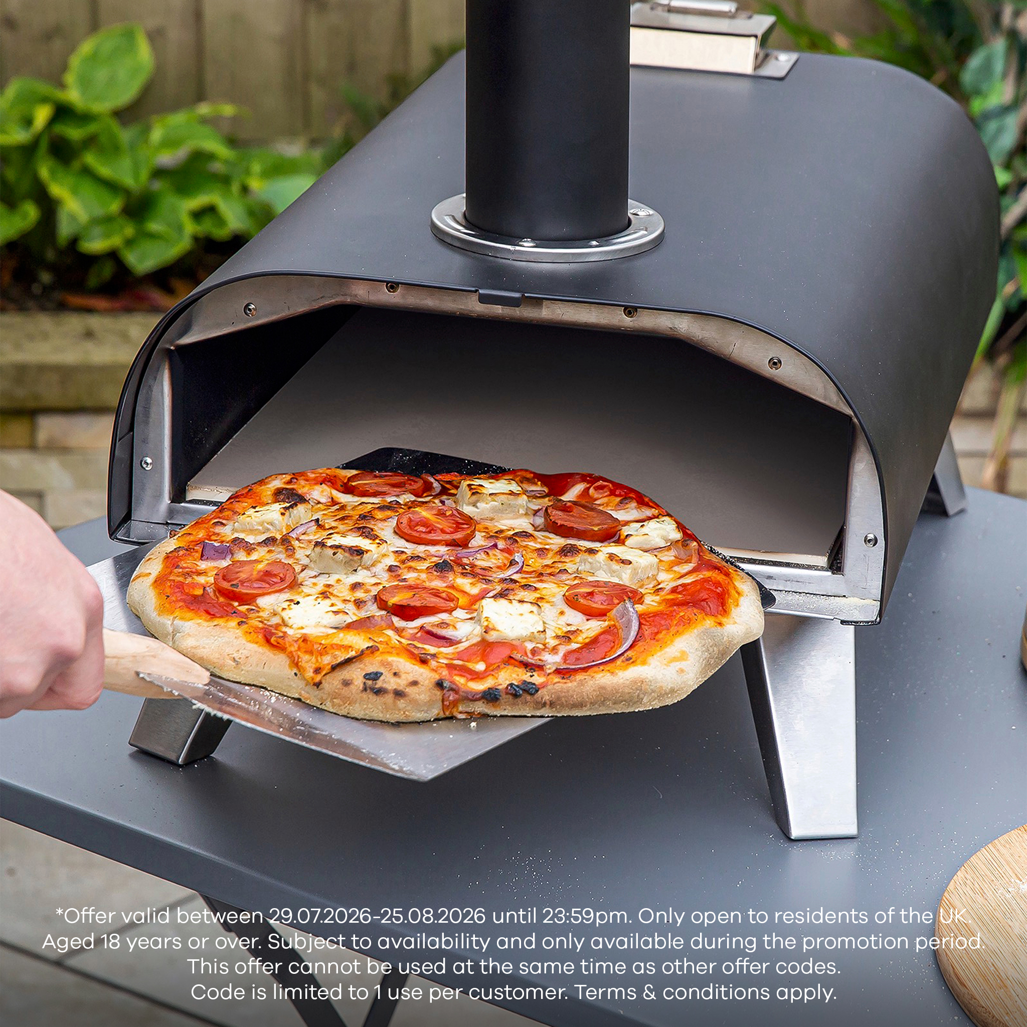 Pizza being removed from a Zanussi outdoor pizza oven using a metal peel, topped with melted cheese, tomato slices, and red onion, on a garden patio table.