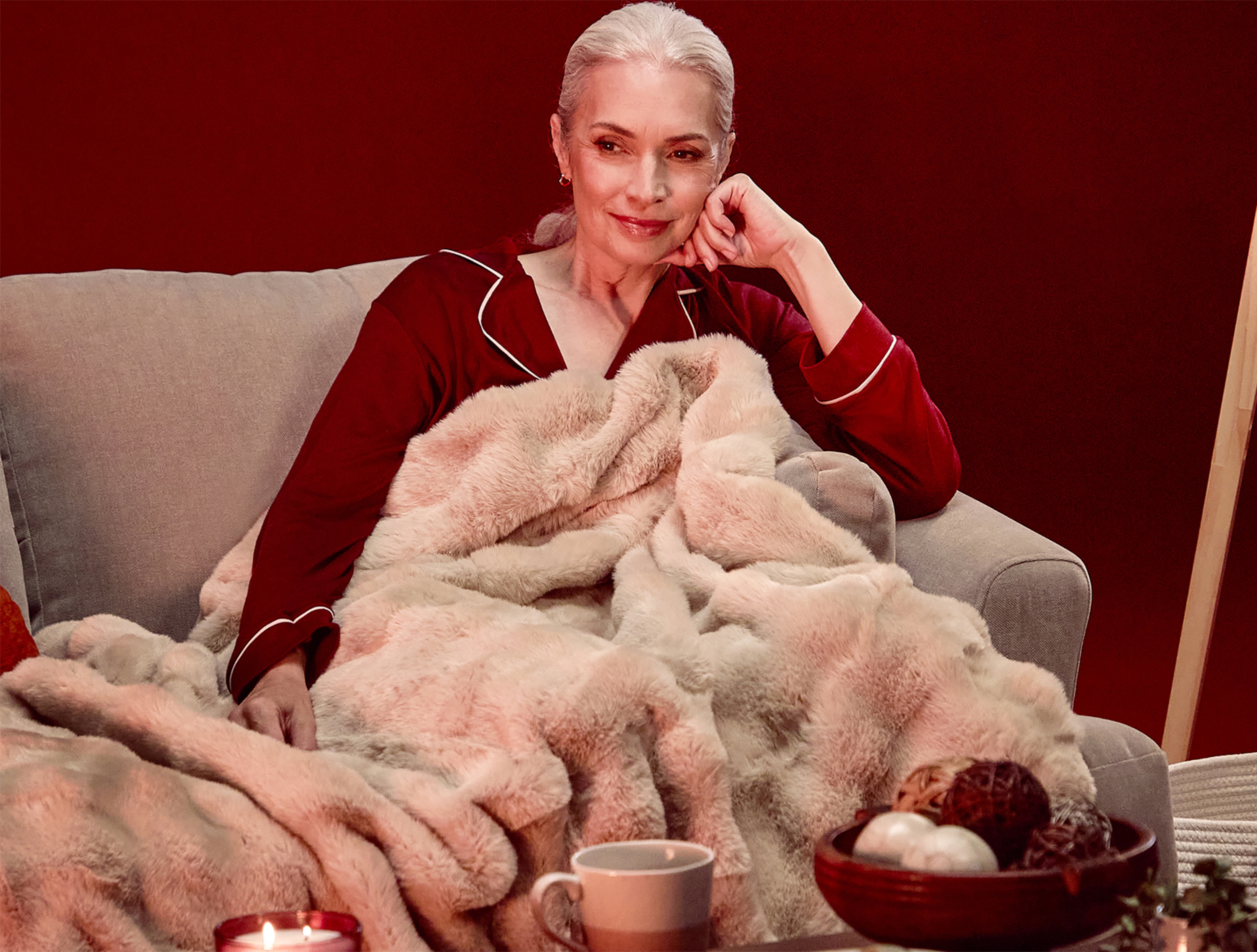 A woman in burgundy pyjamas sat on a sofa under a soft light brown throw.