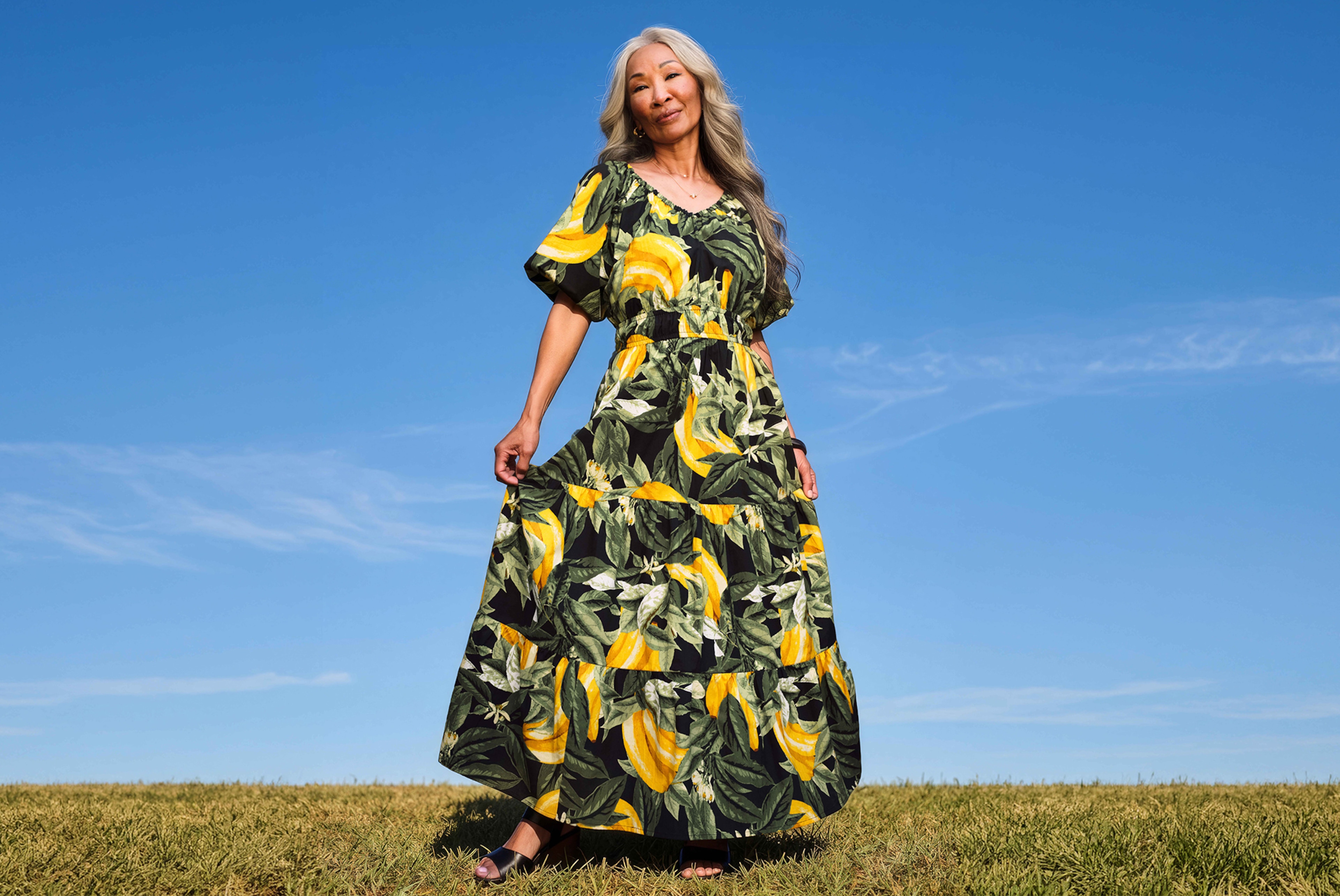 Person wearing a long green dress with yellow leaf pattern standing in a grassy field under a clear blue sky.