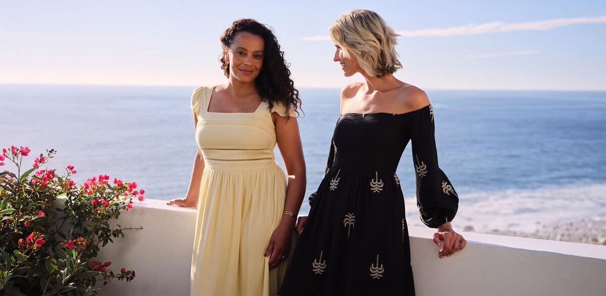 Two women wearing dresses perched on a balcony with the sea in the background.