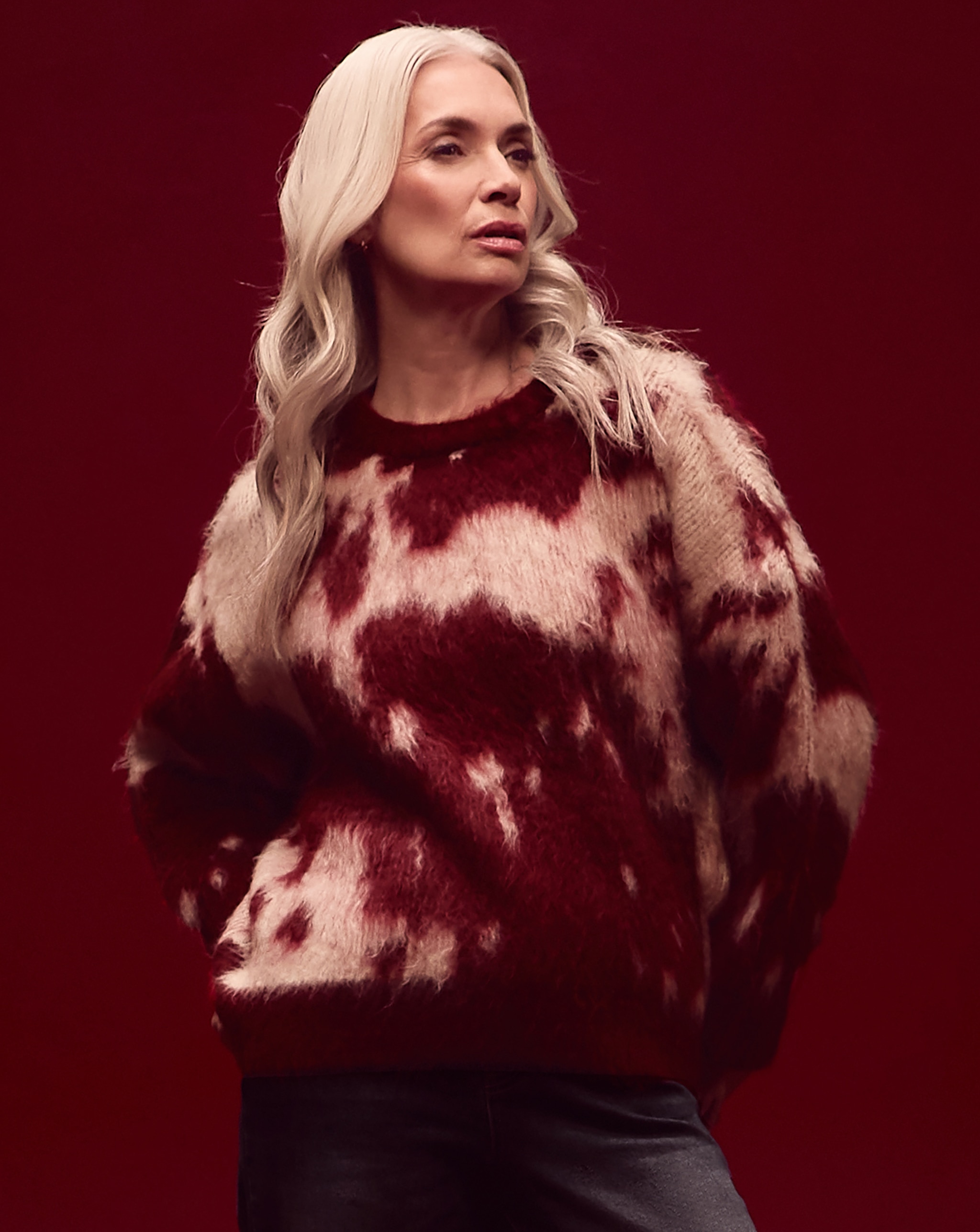 A woman stood up in a red room wearing a red and pink patterned knitted jumper.