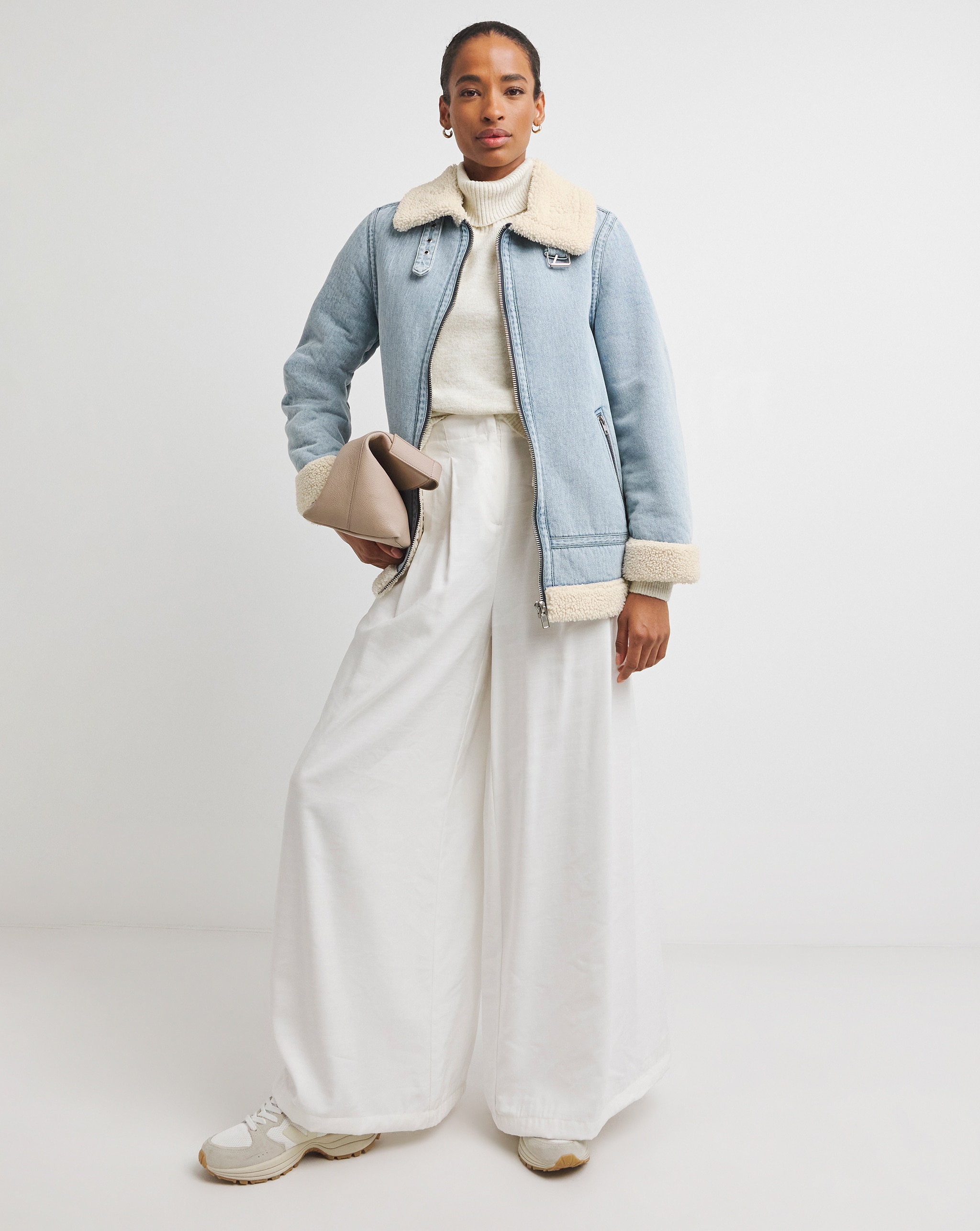 Person wearing a light-wash denim jacket over a cream knit top, styled with wide-leg white trousers, trainers and a neutral clutch bag.