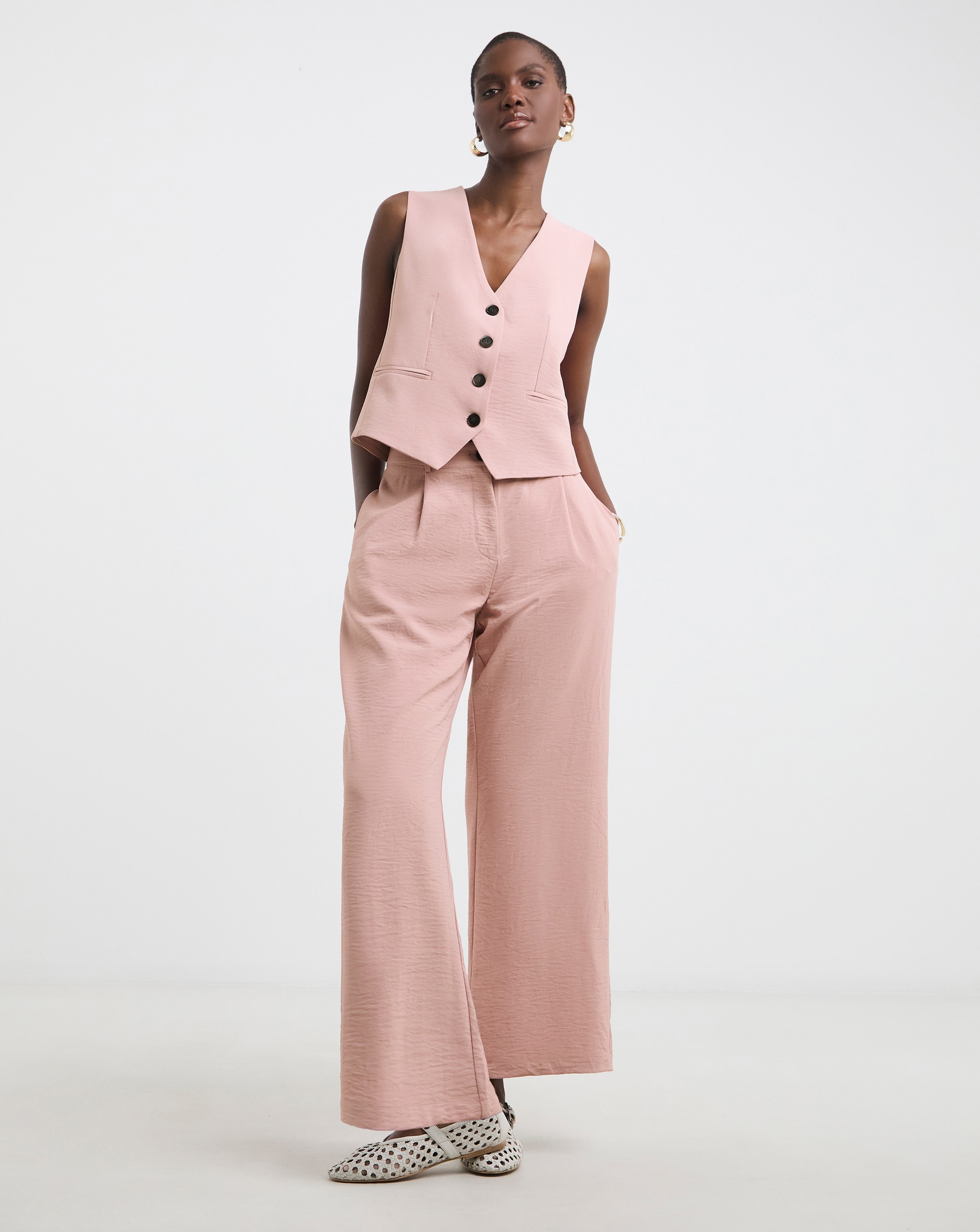 A person standing against a plain white background wearing a light pink sleeveless buttoned vest and matching wide‑leg trousers.