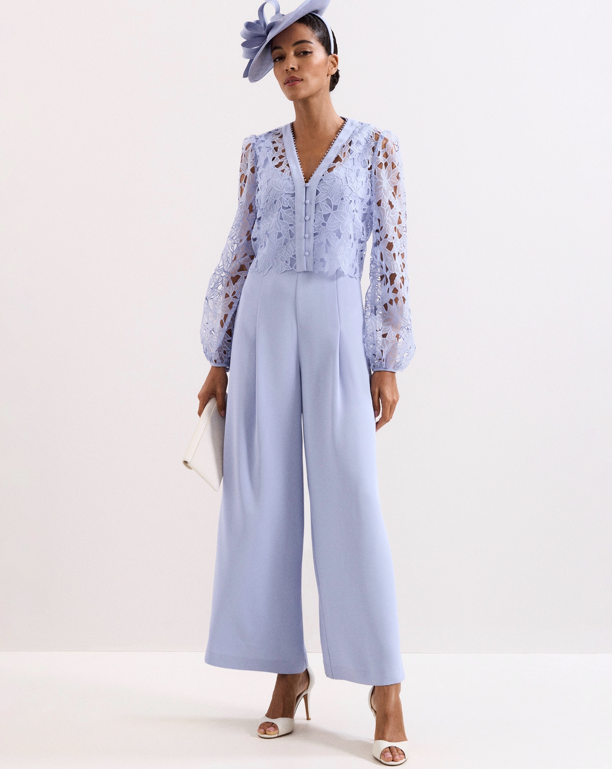 Person wearing a pale blue lace-detail wide-leg jumpsuit with a coordinating fascinator, white clutch bag, and heeled sandals.