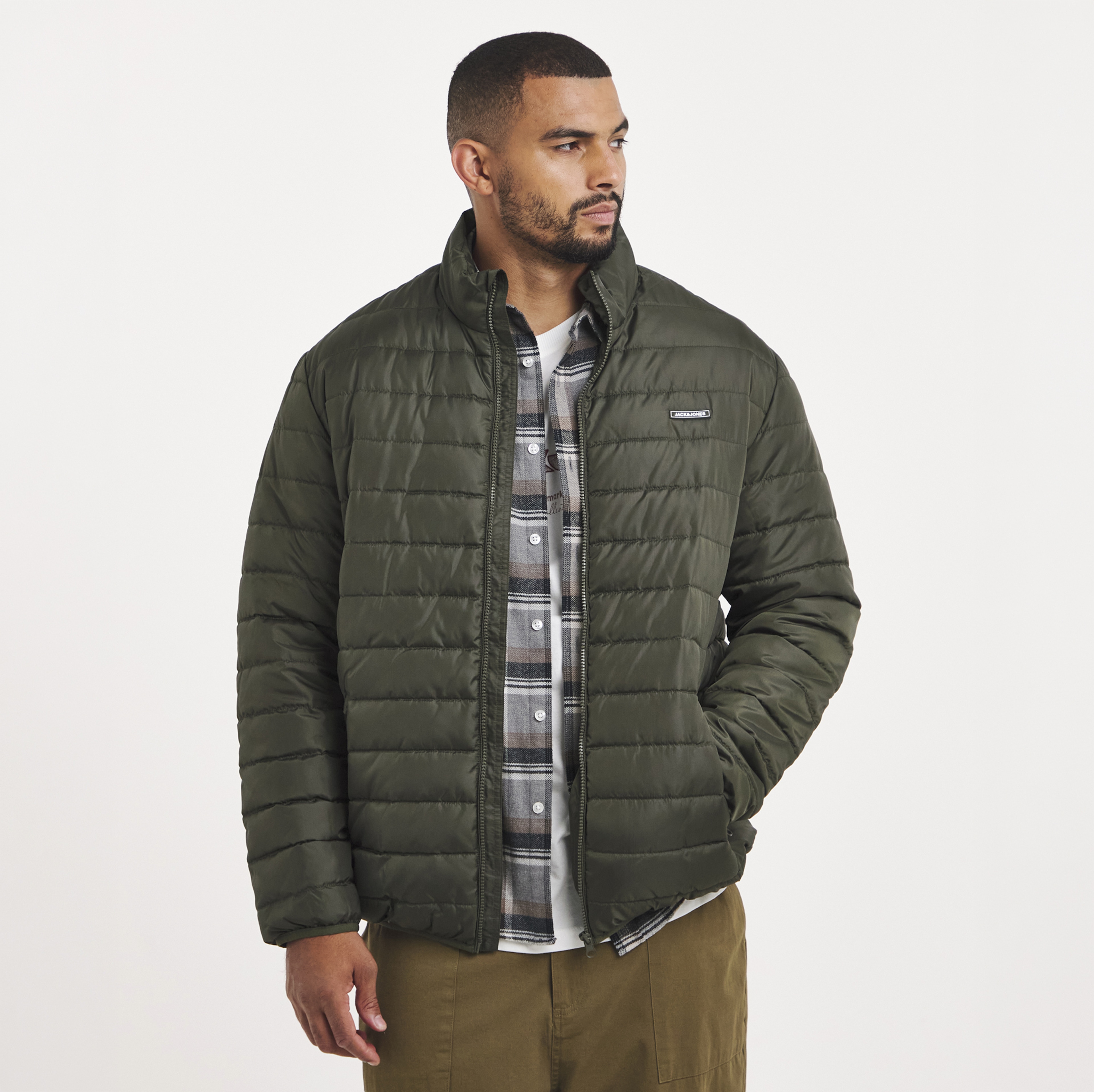 A man wearing a white t-shirt with a grey check shirt and a green quilted jacket on top.
