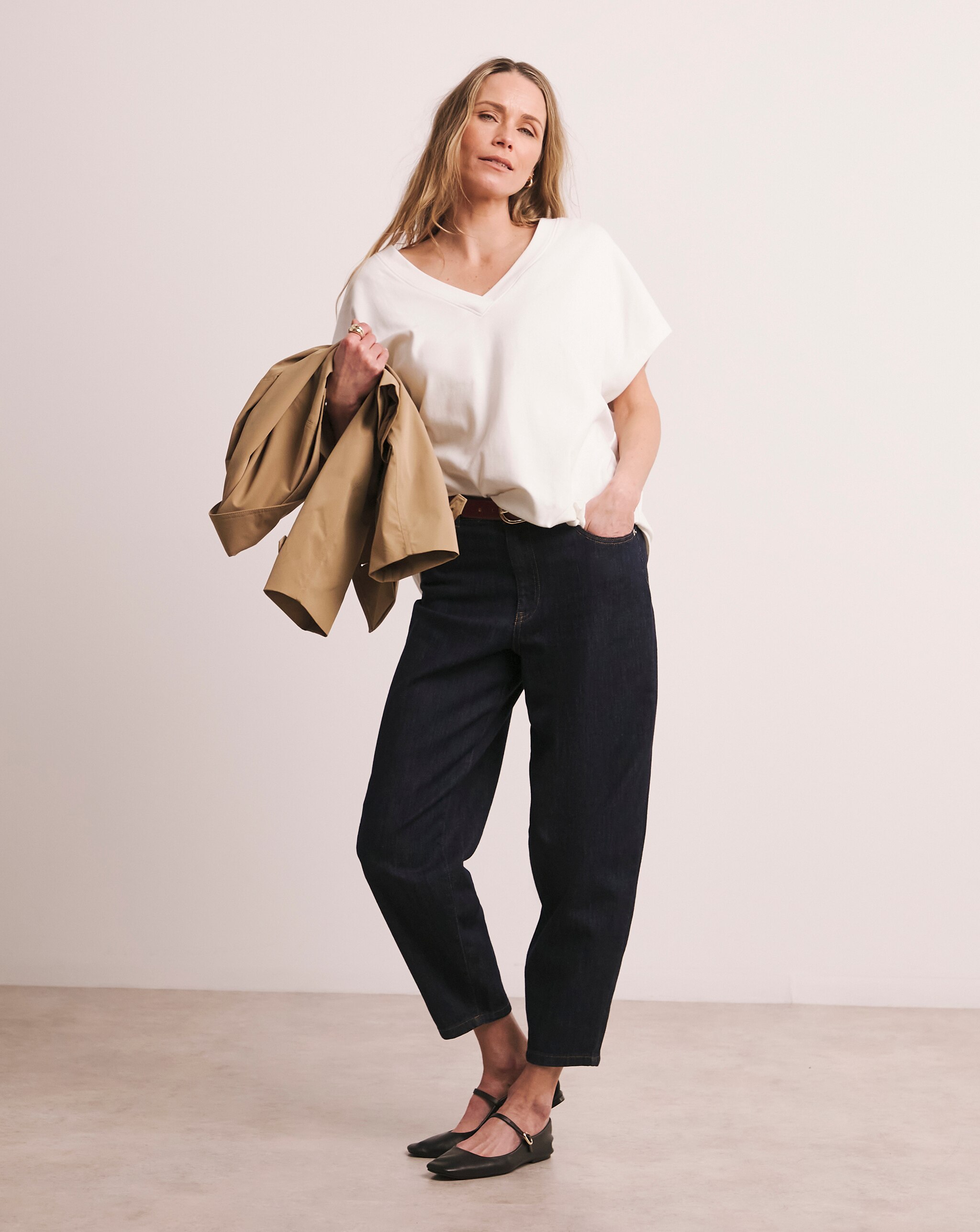 A woman wearing a white v neck tshirt with black jeans holding a coat in her arm.