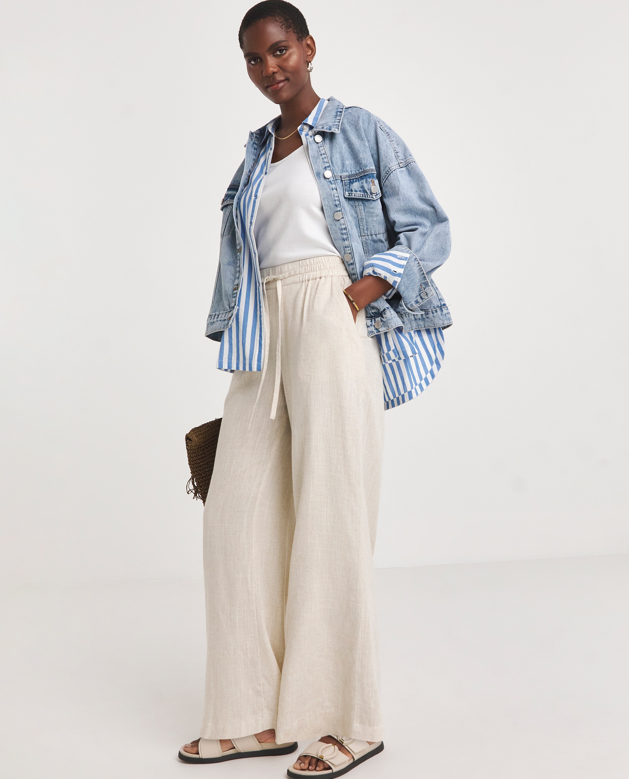 Model is wearing a white top, layered with a stripe shirt and denim jacket over it with a pair of cool stone linen mix wide leg trousers.