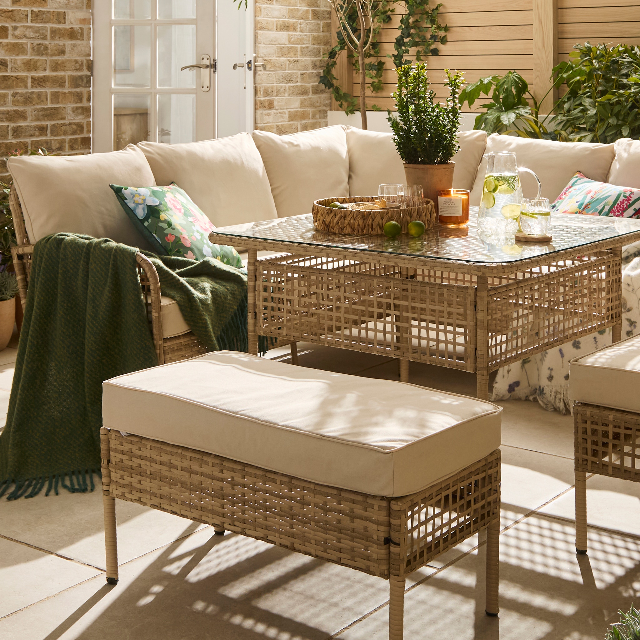 A bright, stylish outdoor living area featuring a cream rattan sofa and matching furniture, including a large ottoman and coffee table. The space is decorated with colourful floral cushions, a green throw, potted plants, fresh limes, drinks and a candle, creating a warm and inviting garden setting.