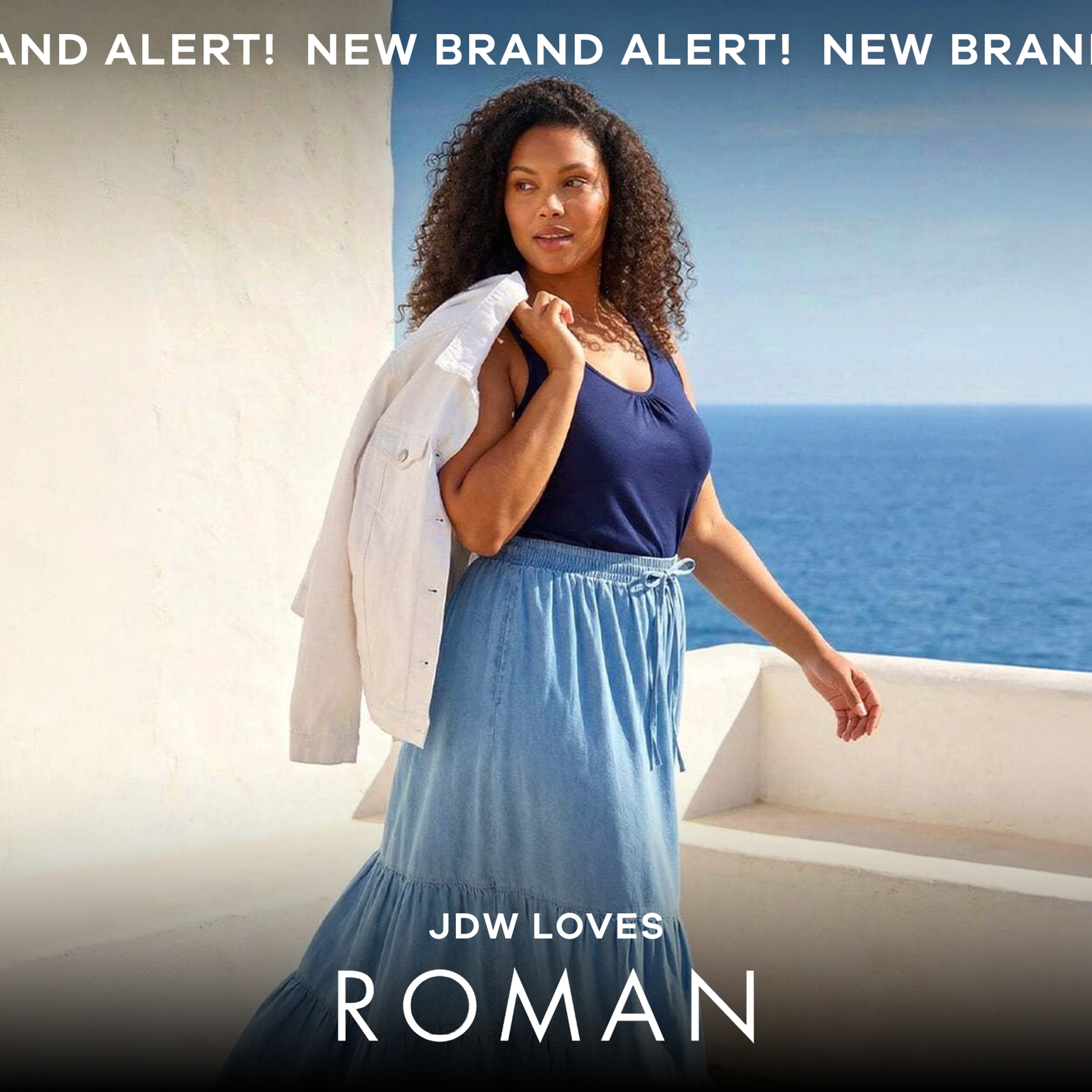 A lady walking outside with the sea in the background wearing a blue top and light blue skirt.