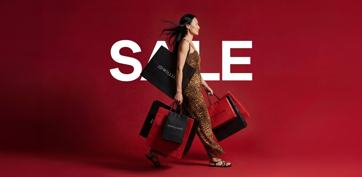 A woman walking with lots of JD Williams shopping bags in front of a red background with the word 'SALE' on it in white