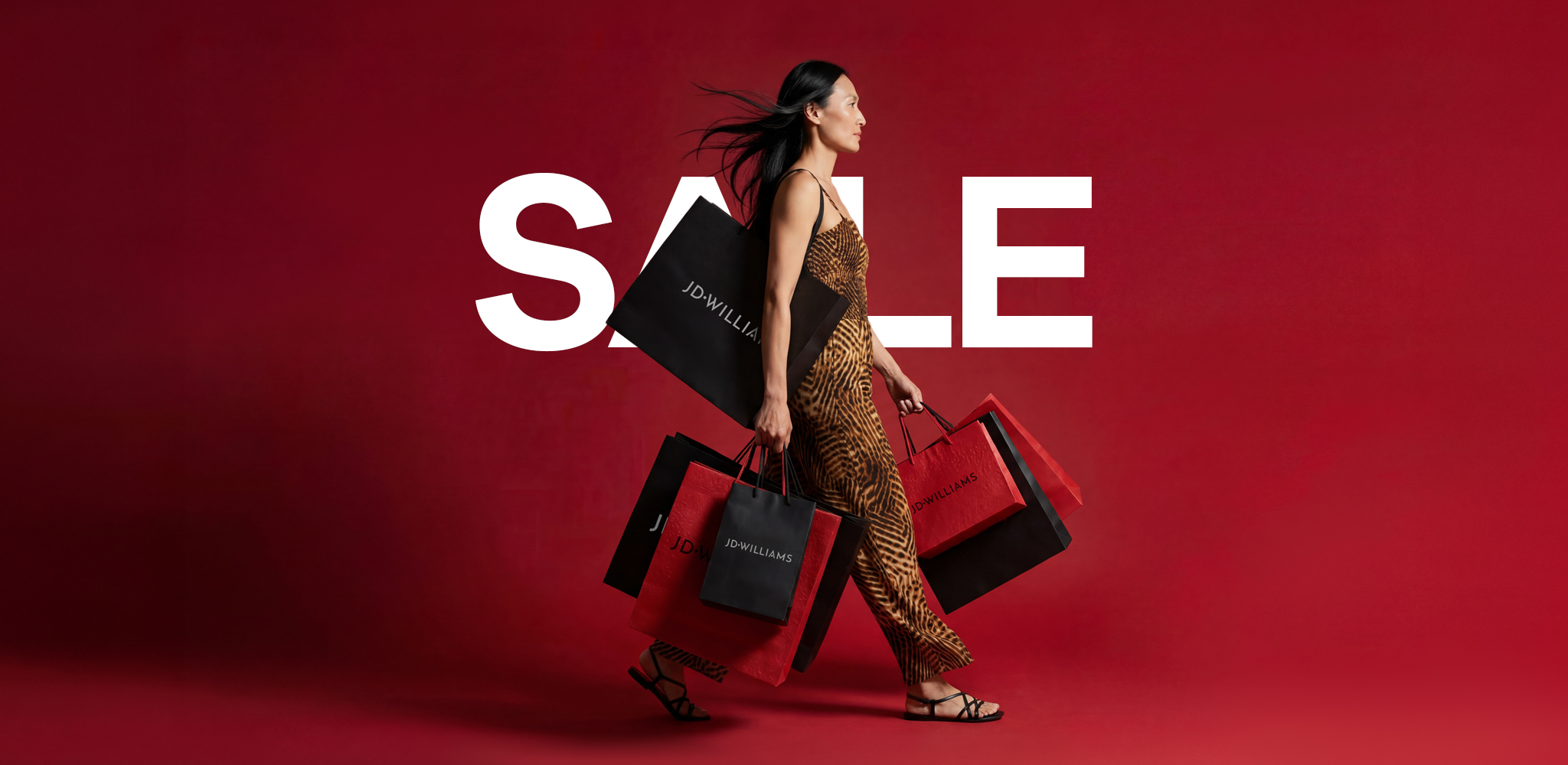 A woman walking with lots of JD Williams shopping bags in front of a red background with the word 'SALE' on it in white