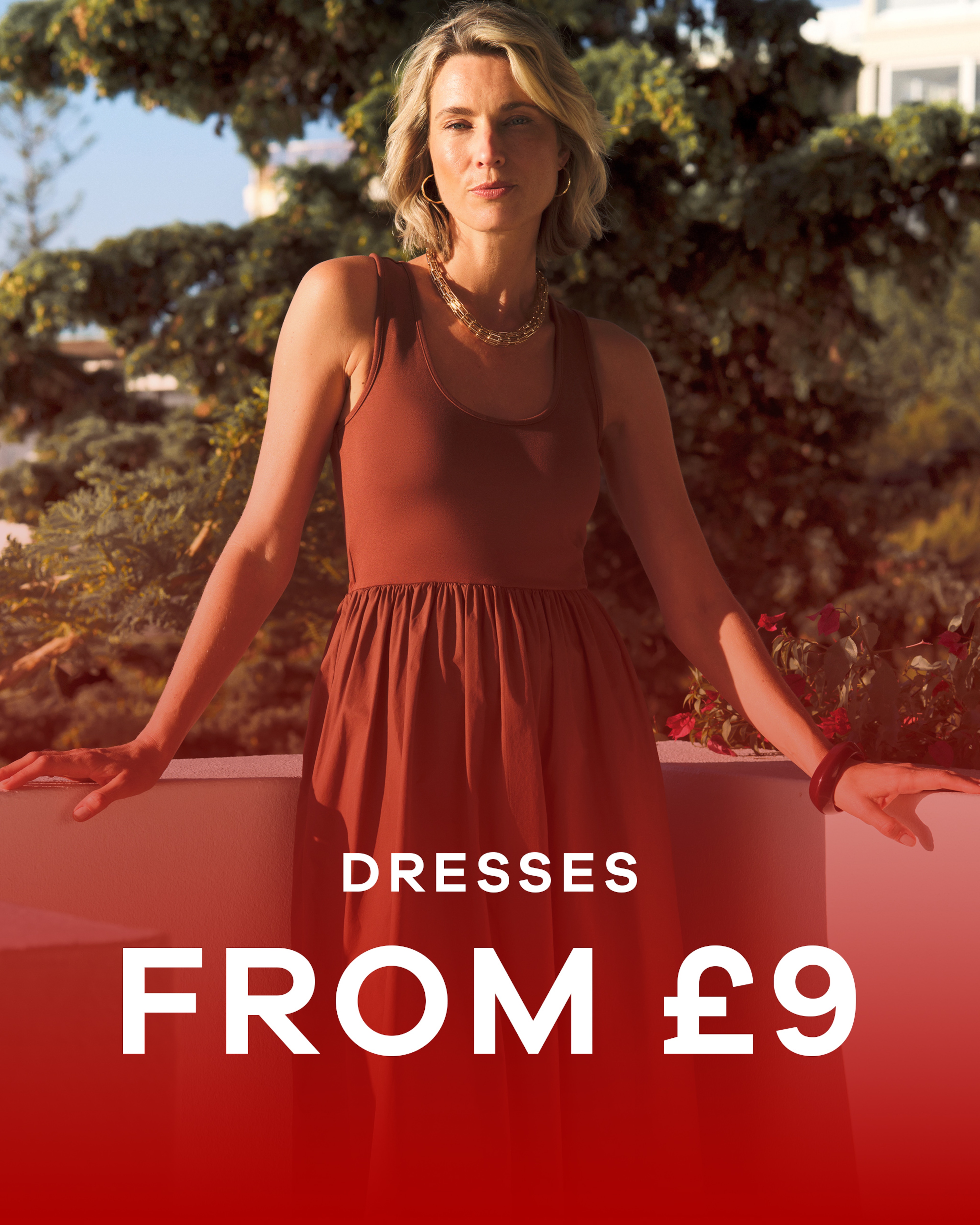 A woman wearing a brown dress on a balcony with greenery behind her in the distance. Dresses from £9