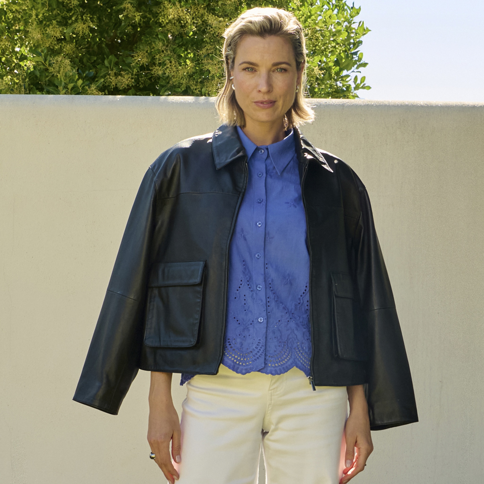 A lady stood in a garden wearing a blue blouse and cream trousers with a jacket draped over her shoulders.