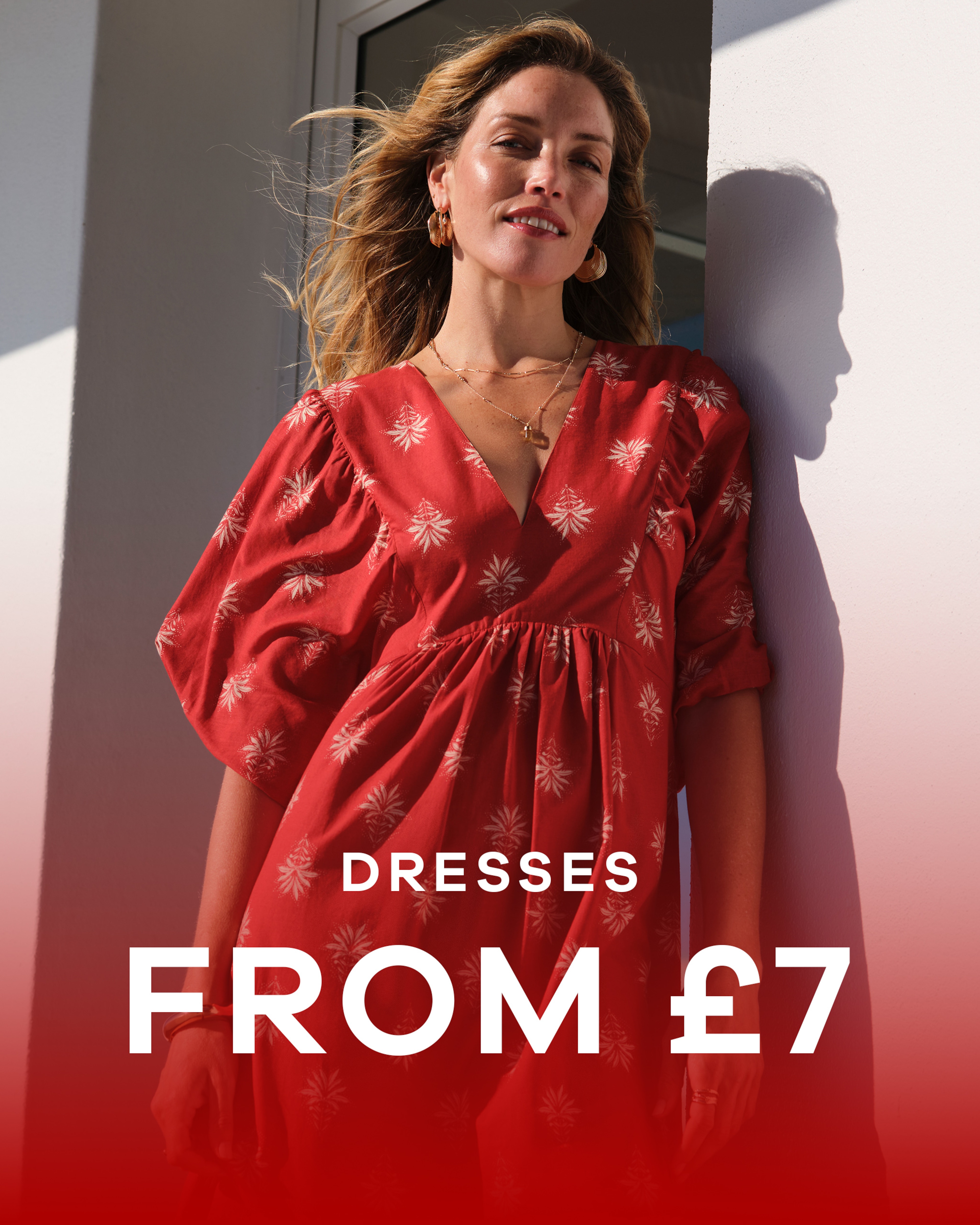 A woman wearing a red print batwing smock dress leaning against a white wall. Dresses from £7.