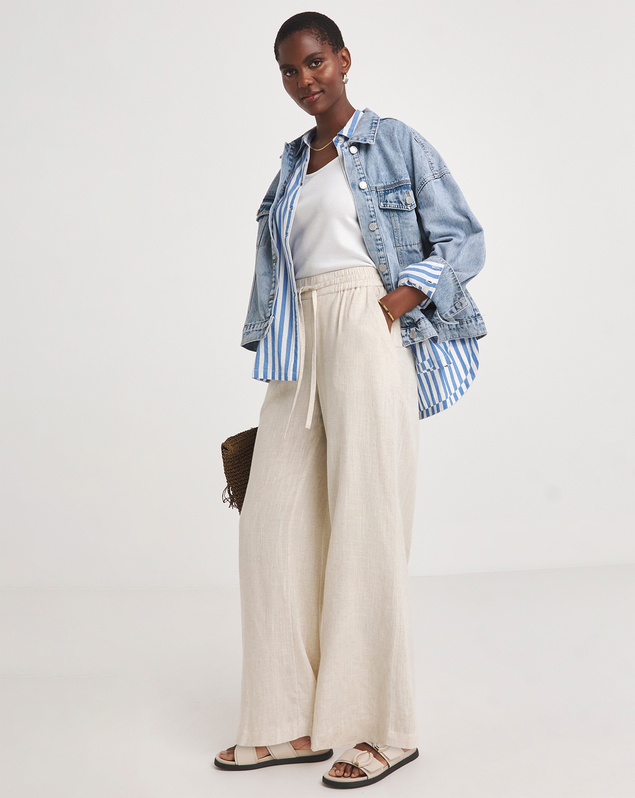 Person wearing a light blue denim jacket over a blue-and-white striped shirt and white vest, paired with wide-leg cream trousers and cream sandals.