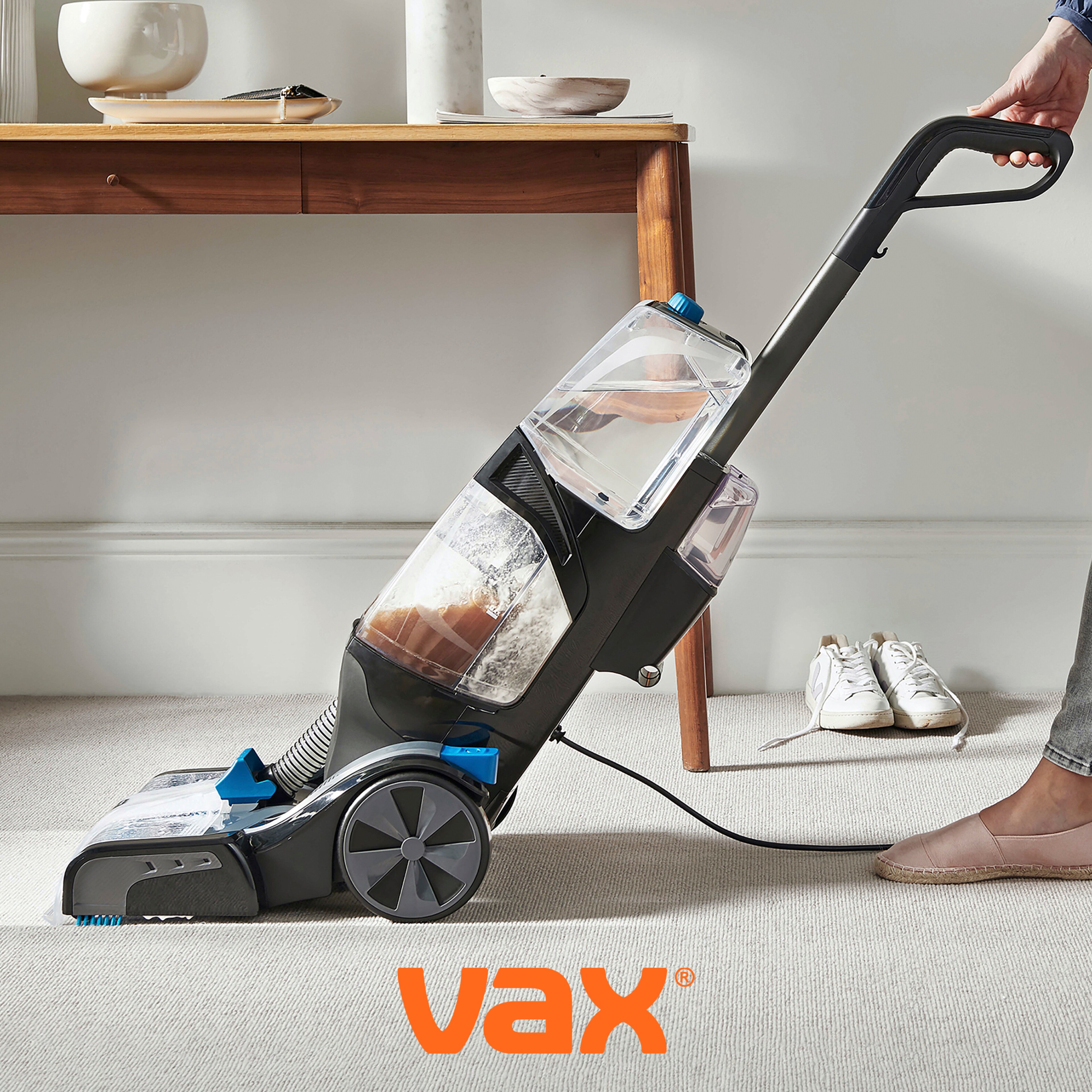 A person using a Vax carpet washer on a light-coloured carpet, with a wooden console table and white trainers in the background.
