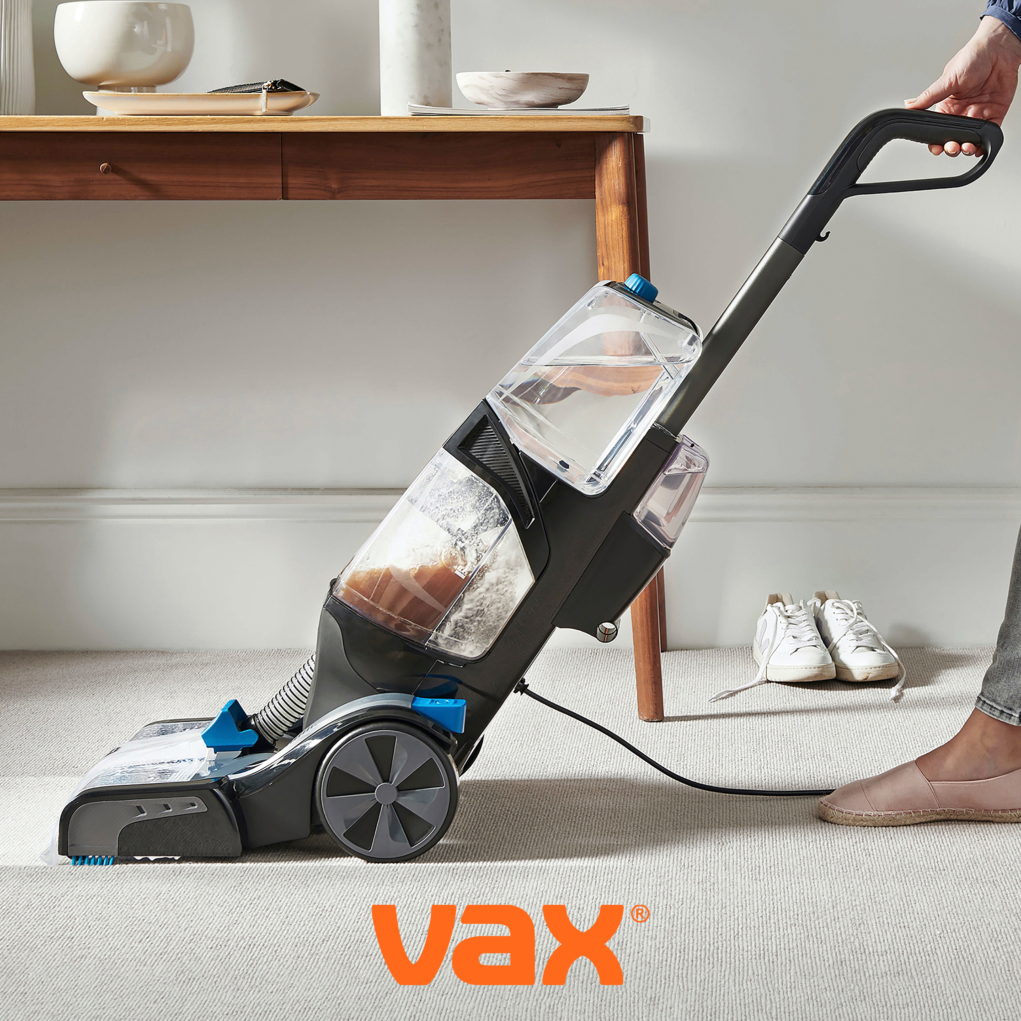 A person using a Vax carpet washer on a light-coloured carpet, with a wooden console table and white trainers in the background.