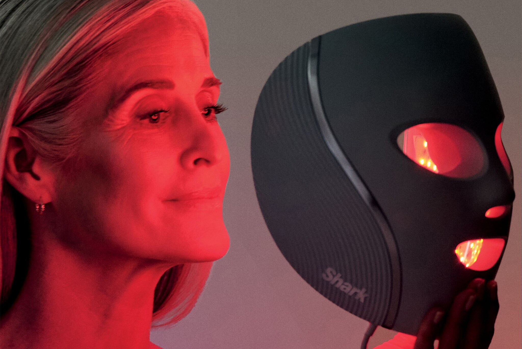A woman illuminated by red light next to a dark grey Shark LED face mask.