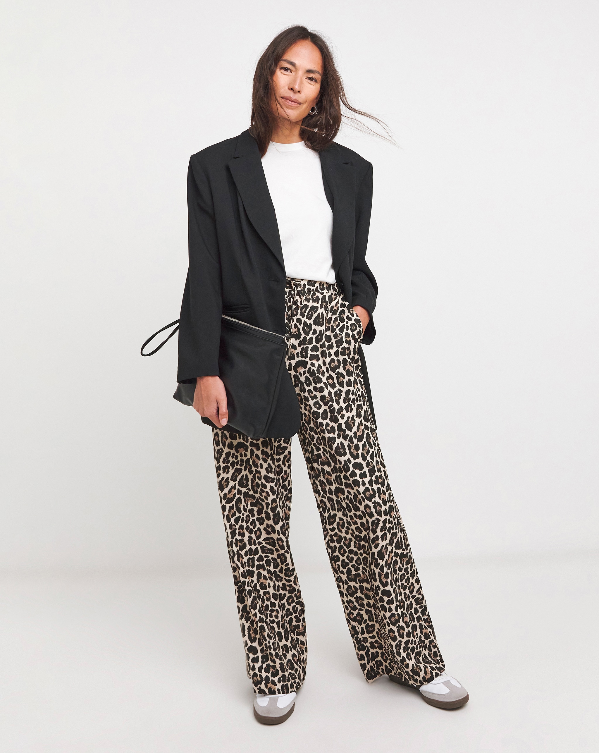 Person wearing leopard-print trousers with a white t-shirt and dark jacket, holding a black bag 
