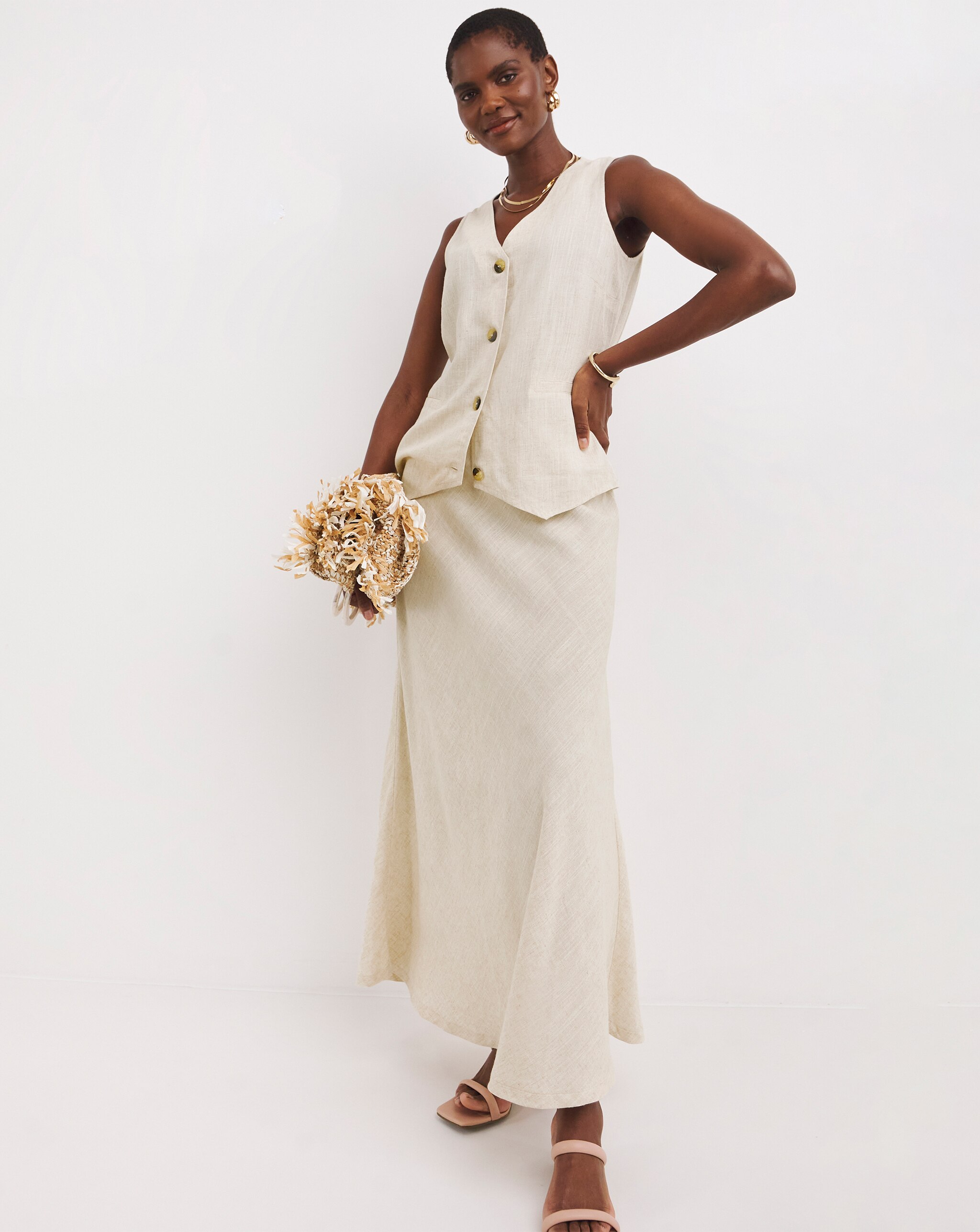 Person wearing a sleeveless natural linen waistcoat and matching maxi skirt, accessorised with gold jewellery and neutral sandals.