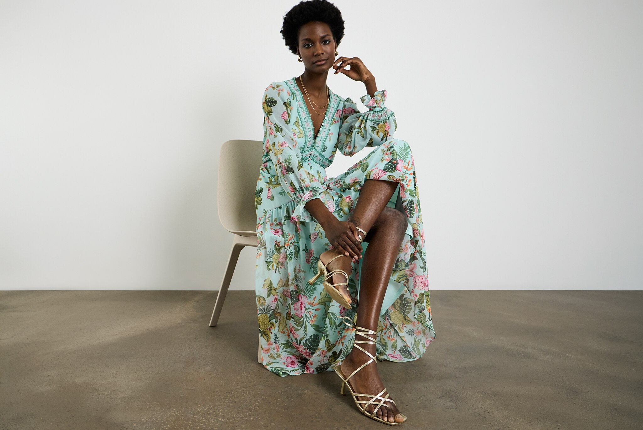 Person seated on a chair wearing a light-blue floral  Joanna Hope dress and lace-up heeled sandals, posed in a studio with a plain grey backdrop and polished concrete floor.