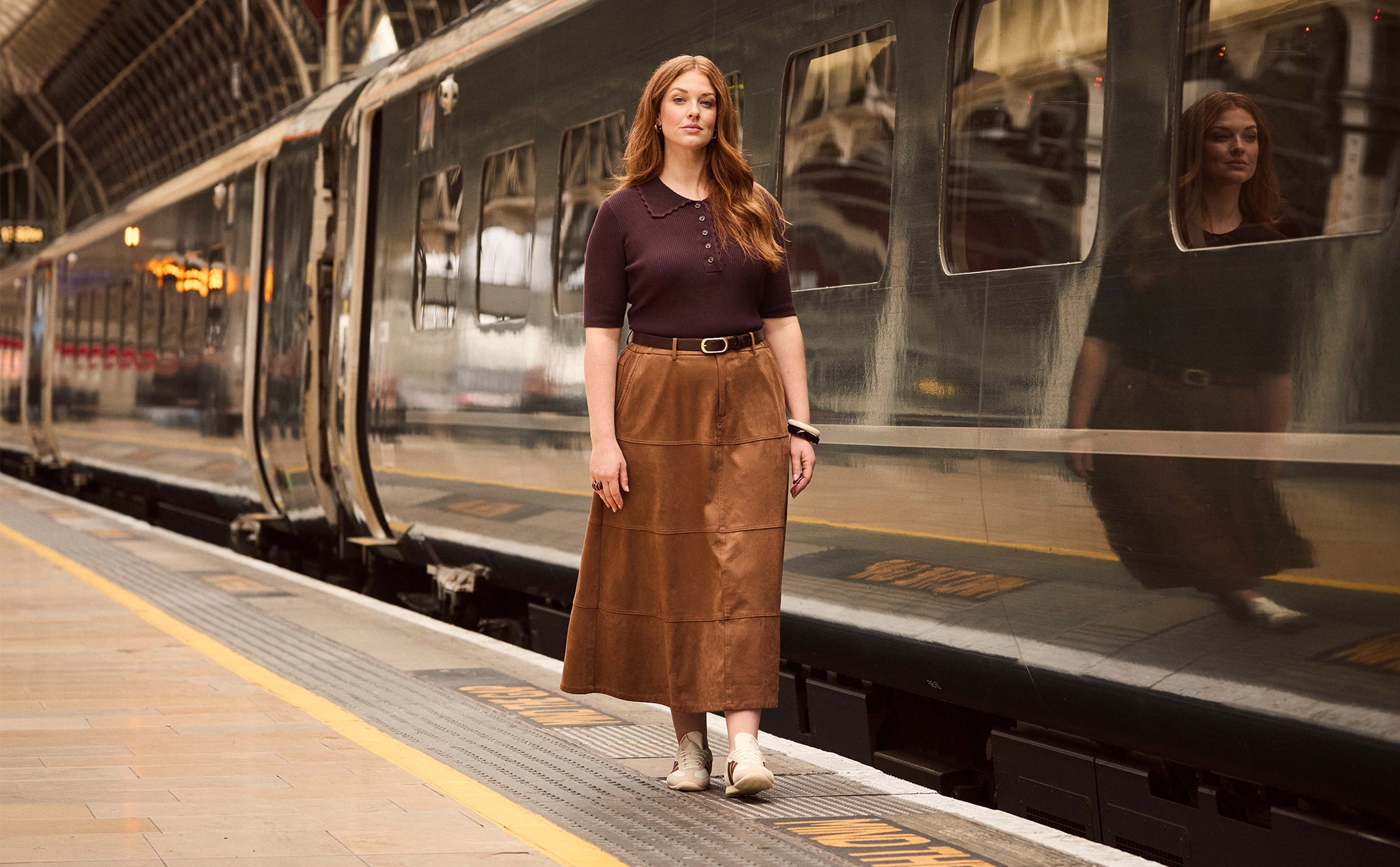 A lady wearing a burgundy top and brown suede maxi skirt walking next to a train at a station.
