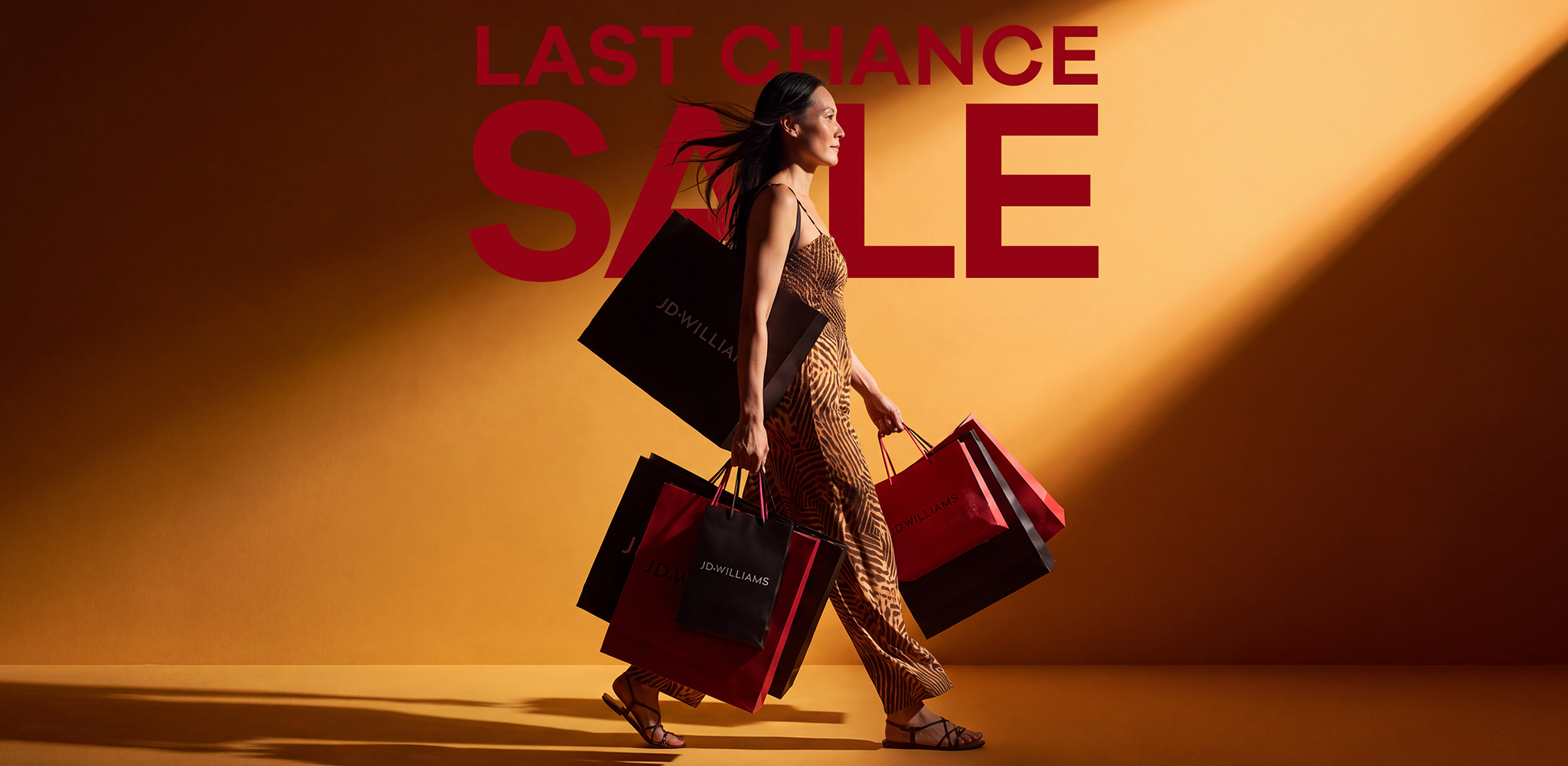 A woman walking with lots of JD Williams shopping bags in front of a orangey yellow background with the word 'SALE' on it in white
