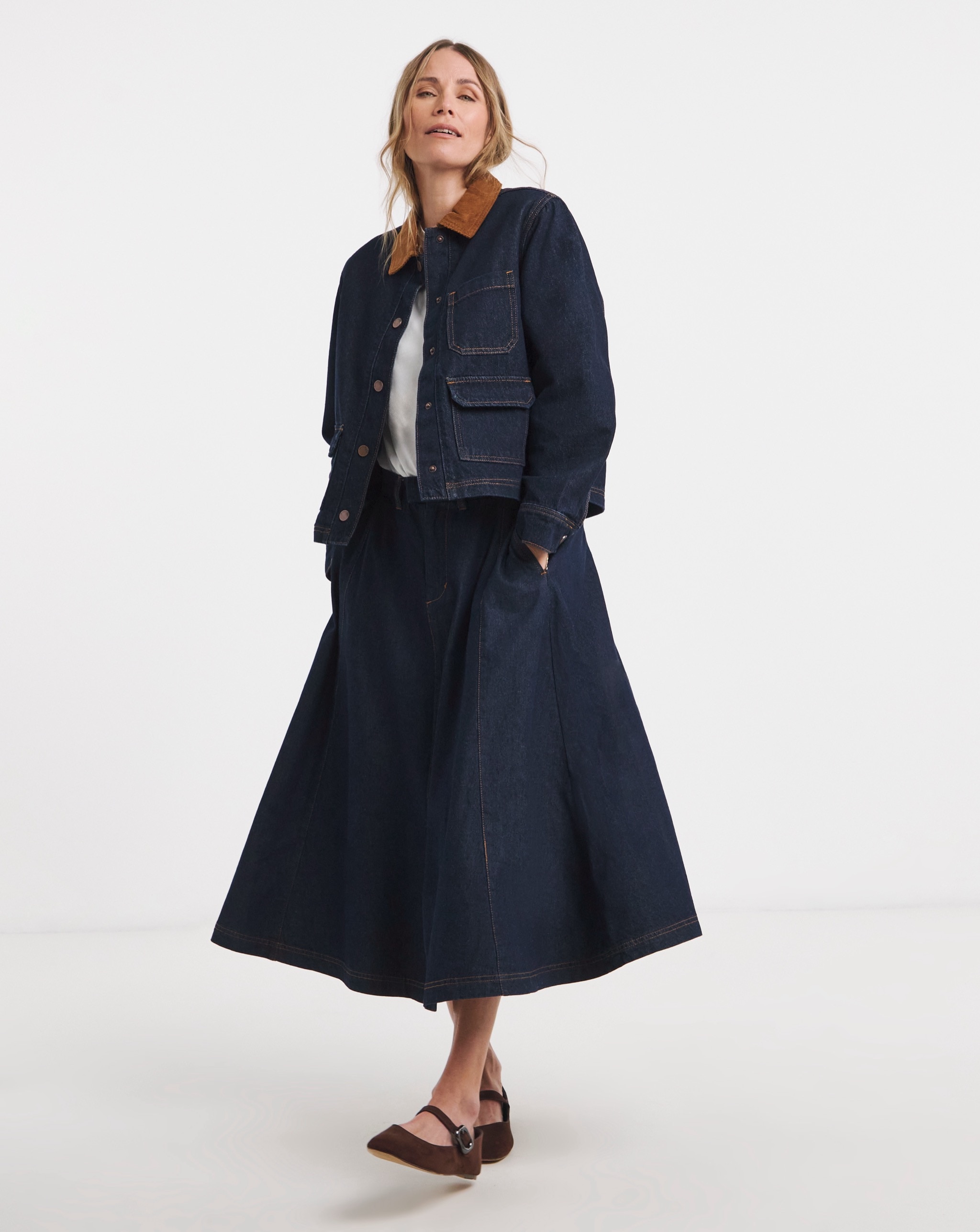 Person wearing a dark indigo denim jacket, styled with a matching midi skirt, white top and brown flats.
