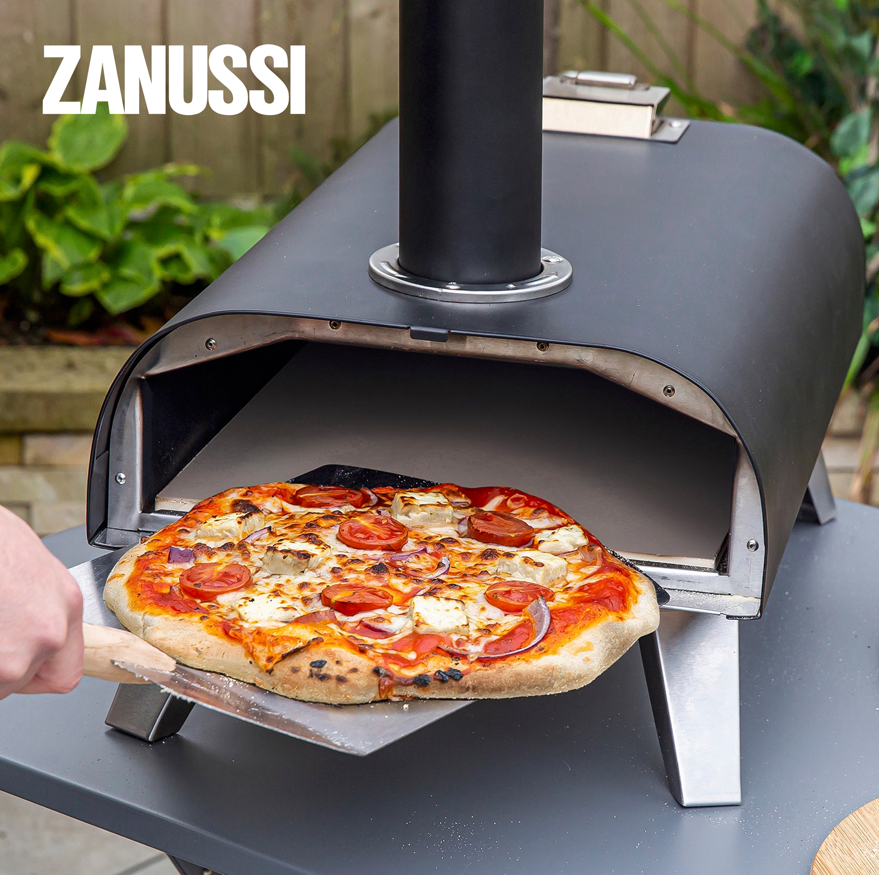 Outdoor pizza oven with a chimney, a freshly baked pizza being slid out on a metal peel.