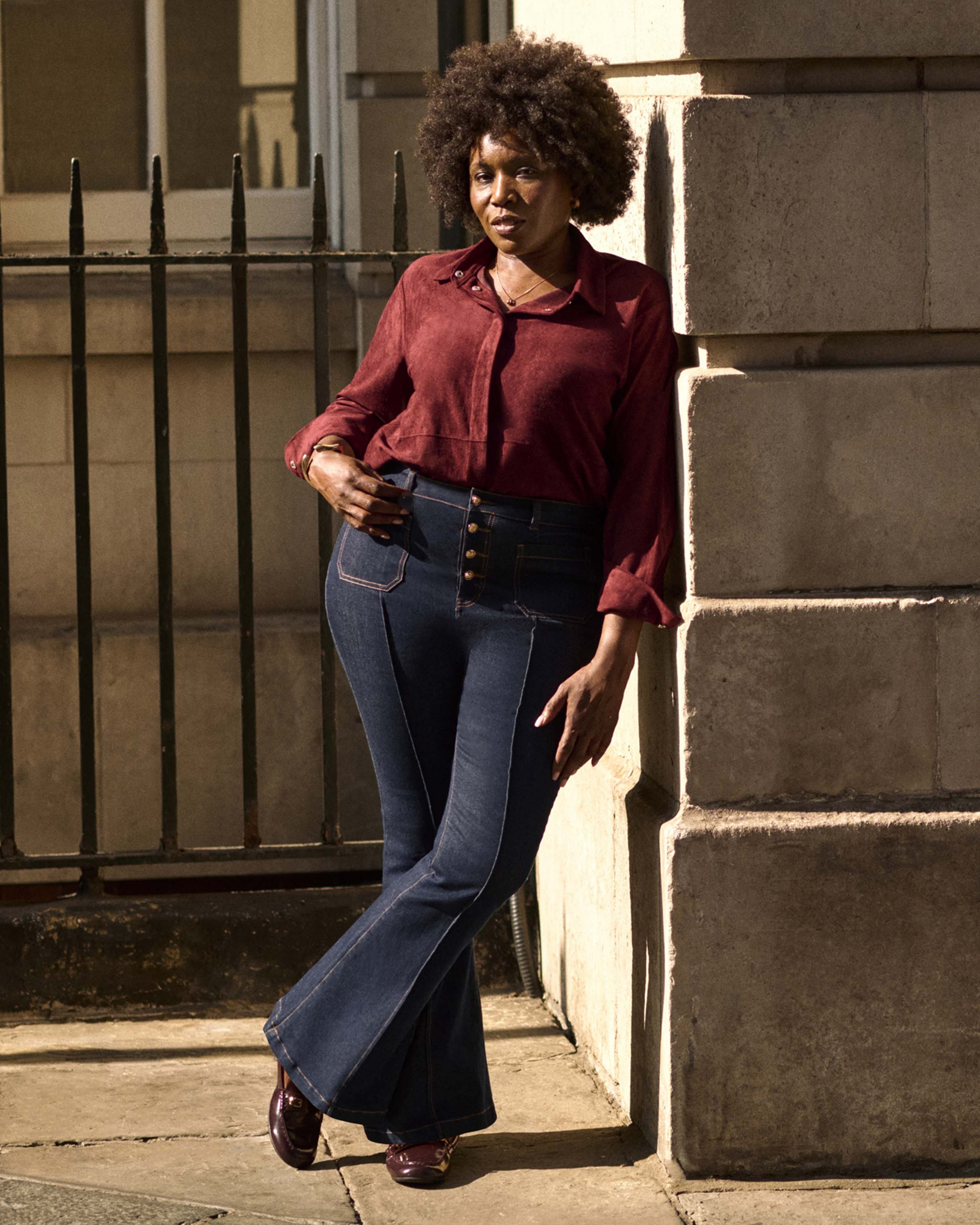 A woman stood leaning against a wall wearing a maroon top and indigo jeans.