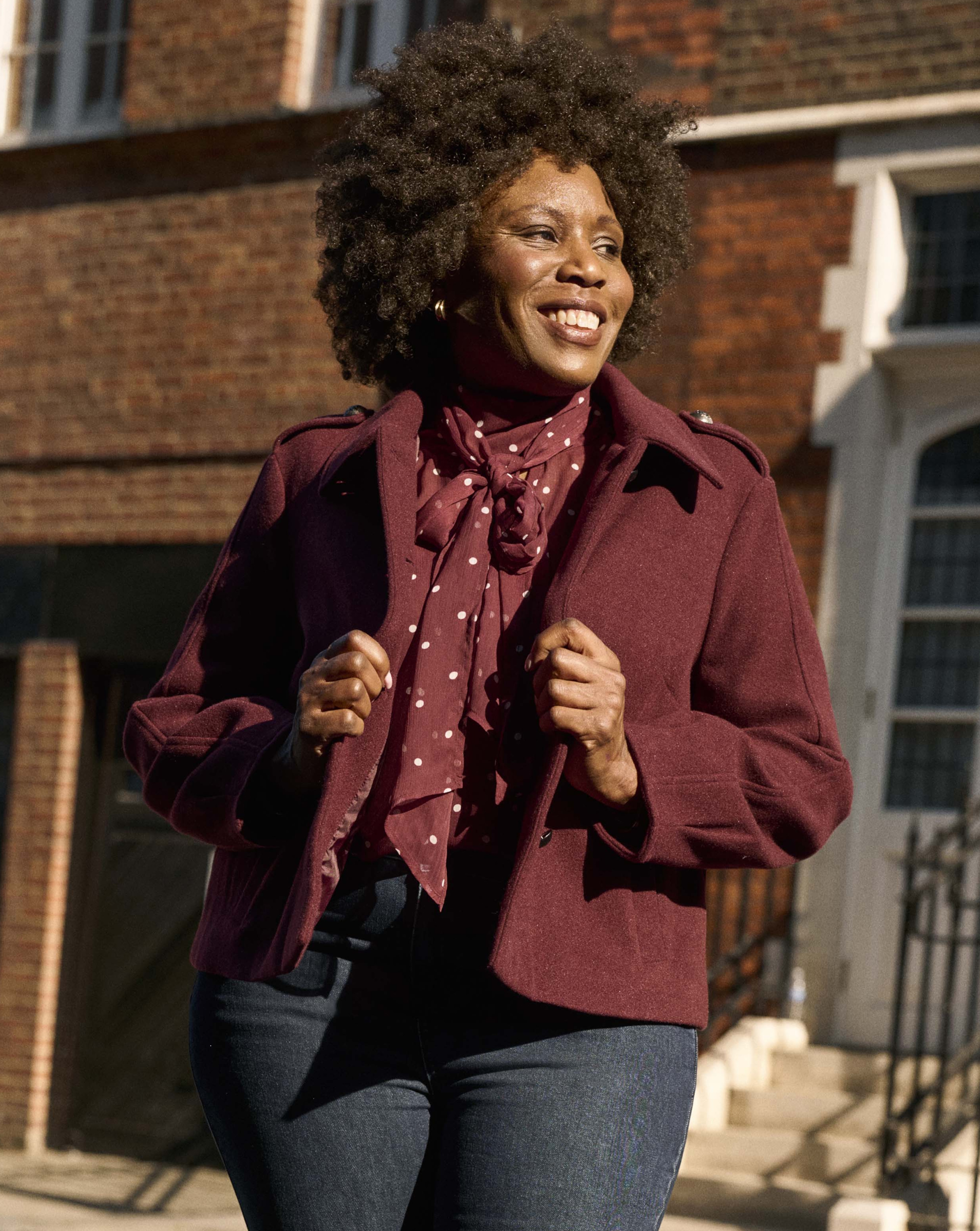 A woman stood outside wearing a burgundy blouse with white spots, a burgundy jacket and blue jeans.