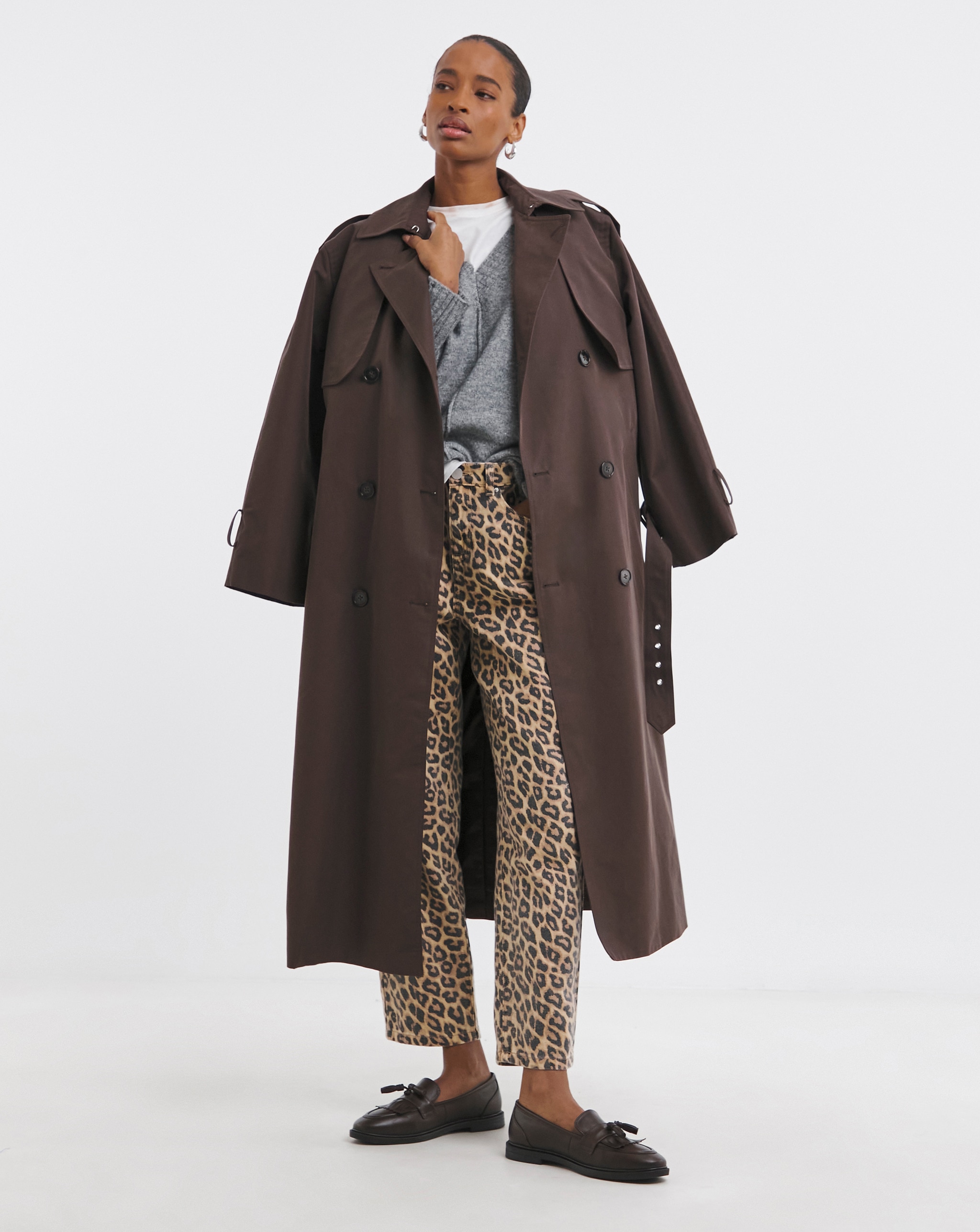 Person wearing a long brown trench coat over a grey jumper and leopard-print trousers with black loafers.