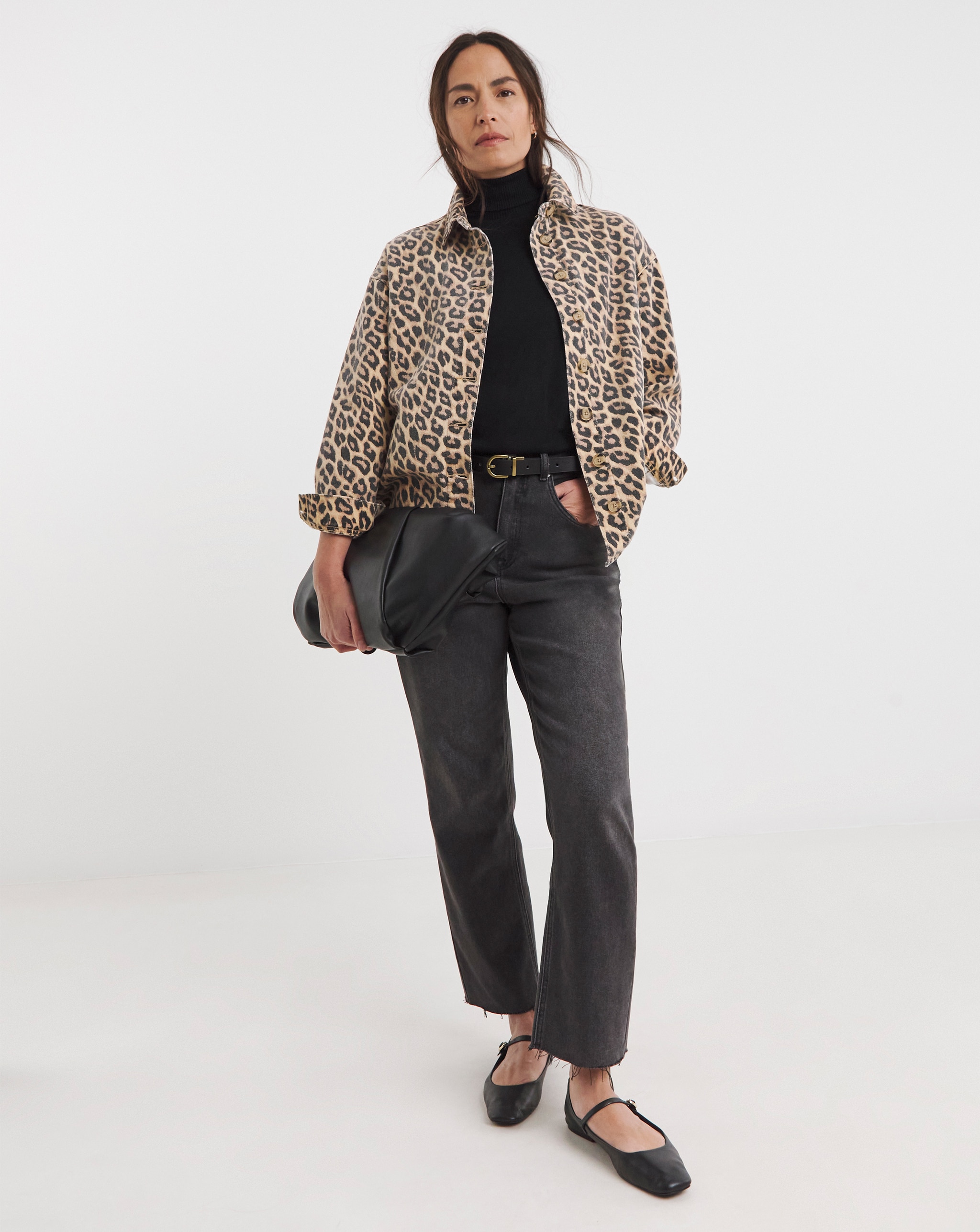Person wearing a leopard-print denim jacket over a black roll-neck top, styled with charcoal jeans, black flats and a black clutch bag.