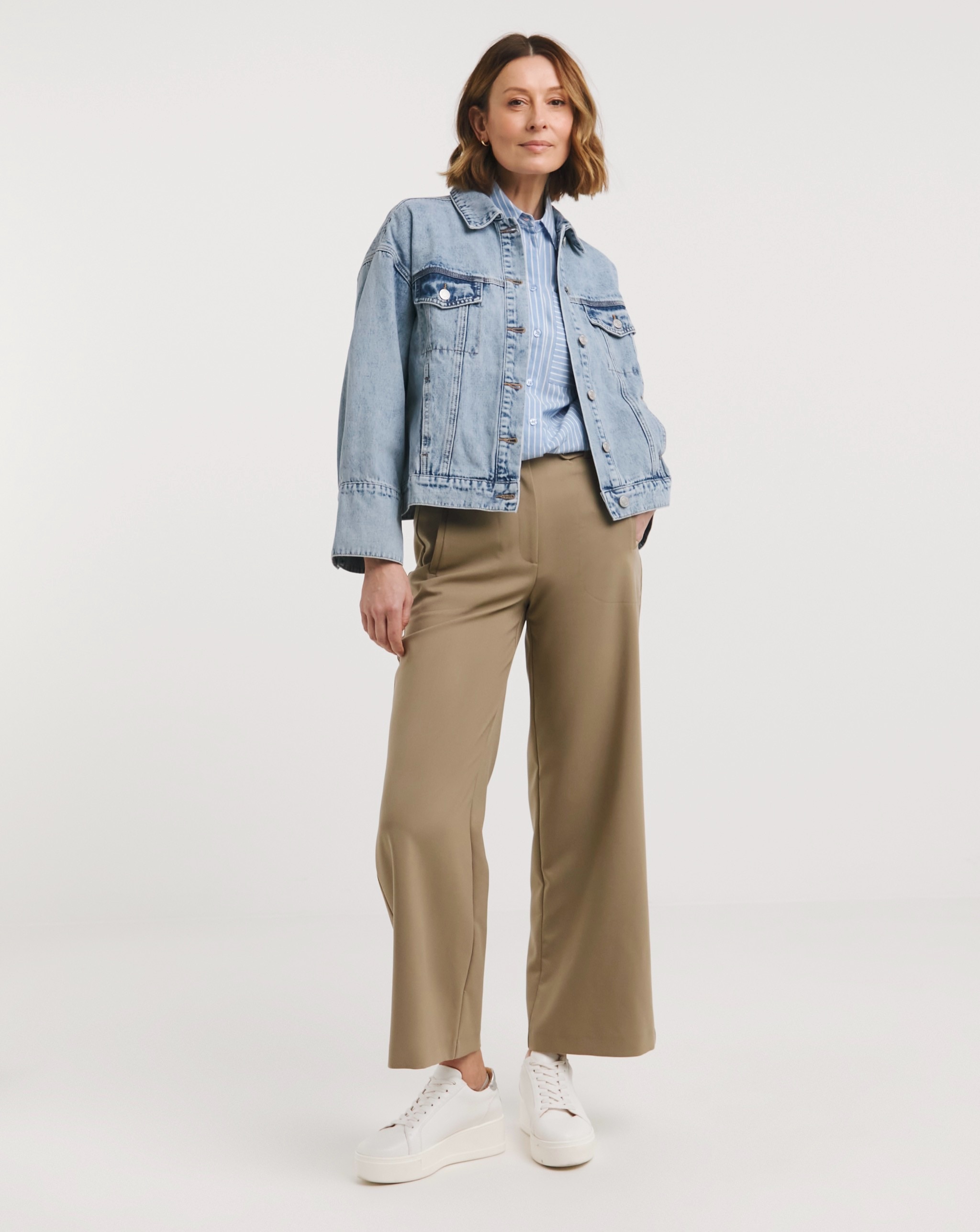 Person wearing a light-wash denim jacket over a blue striped shirt, styled with taupe wide-leg trousers and white trainers.