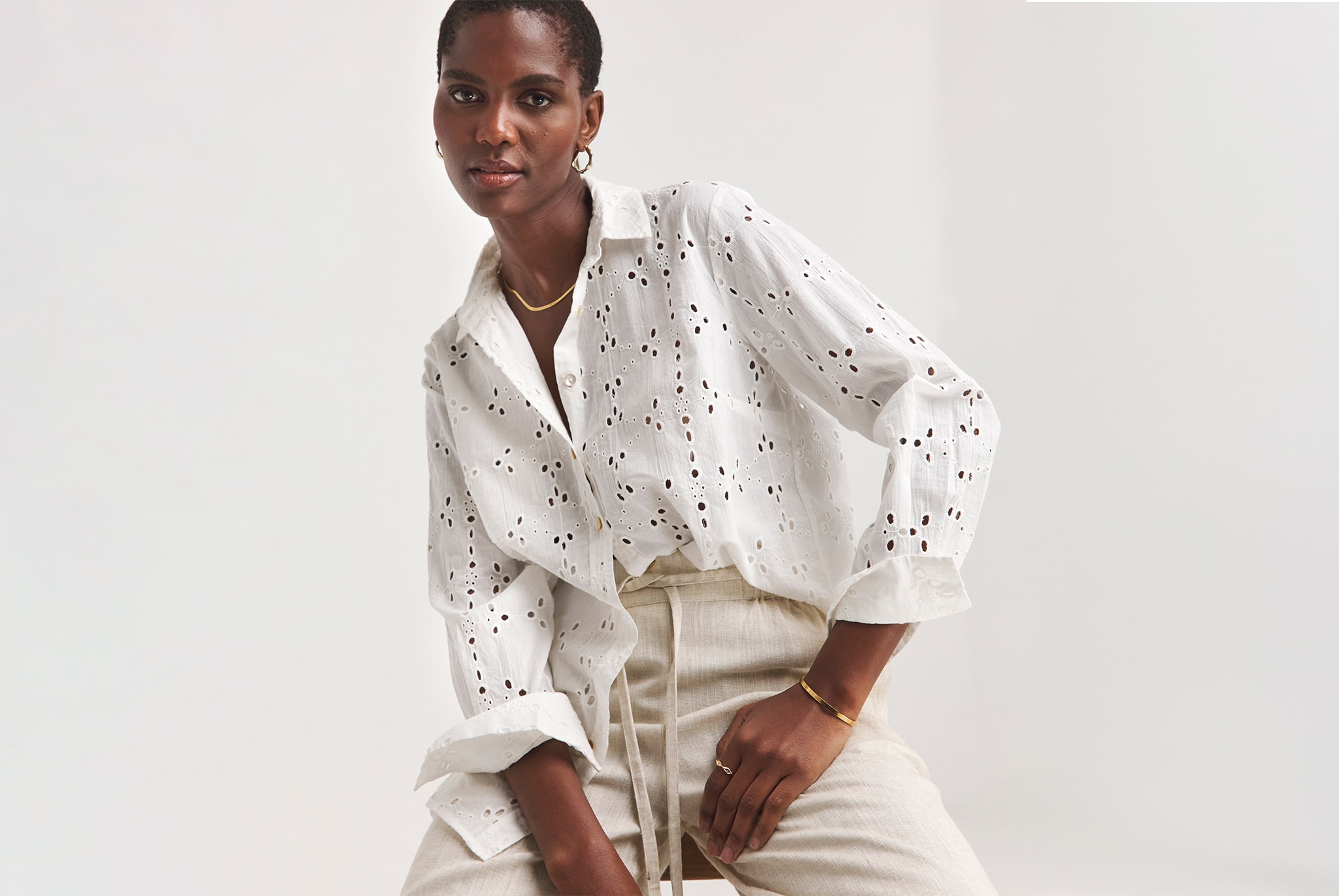 Person seated in a studio setting, wearing a white eyelet embroidered button-up shirt with rolled sleeves, beige drawstring trousers, and gold jewellery.