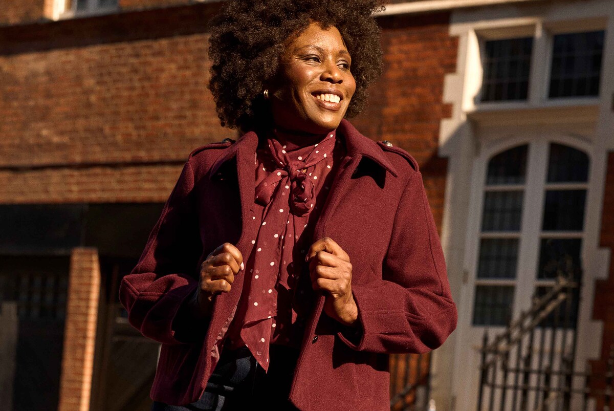 Person wearing a burgundy wool coat layered over a polka-dot blouse with tie neck, styled outdoors in warm sunlight.