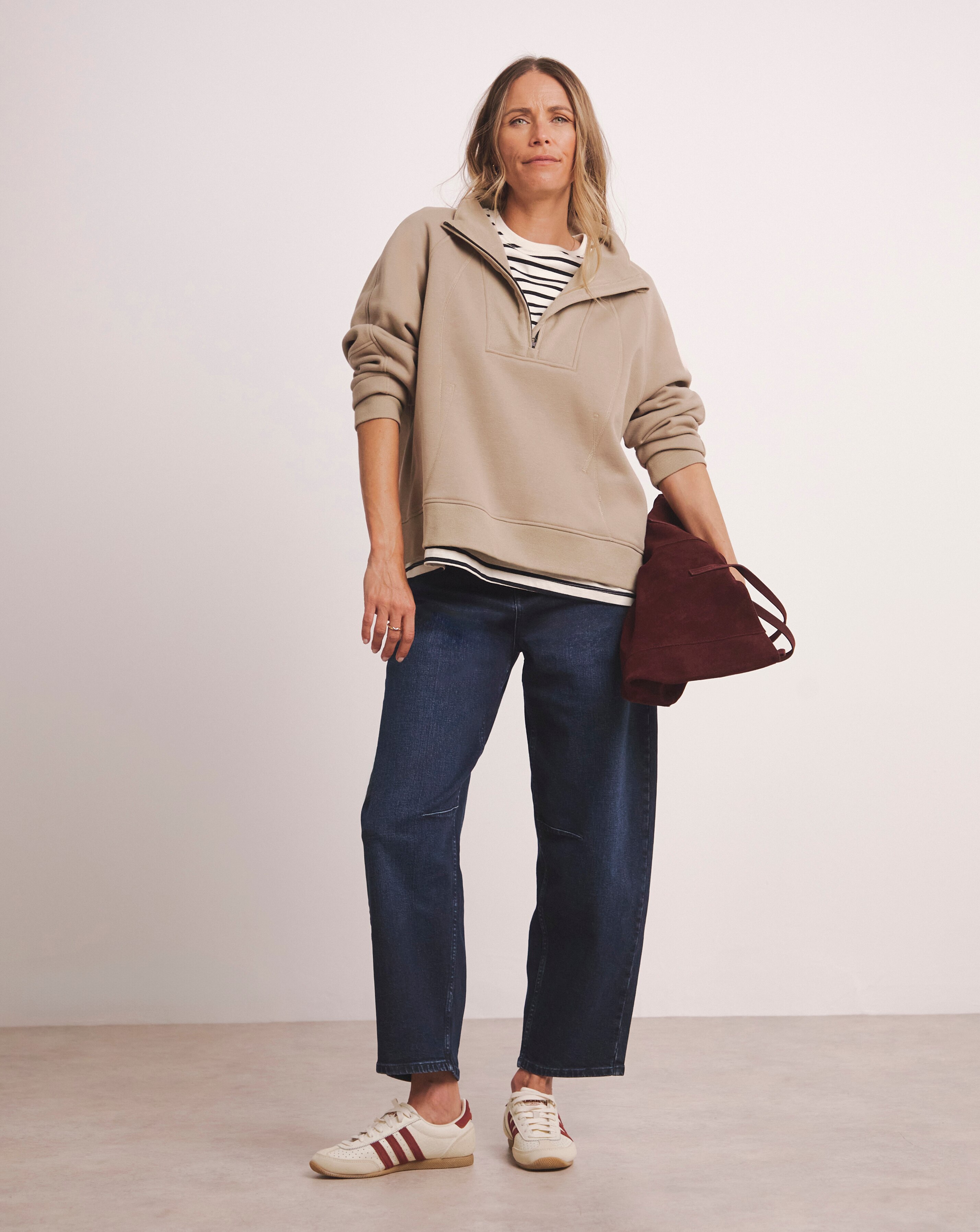 A woman wearing a quarter zip stone coloured top with a white and blue striped top underneath with dark blue jeans.