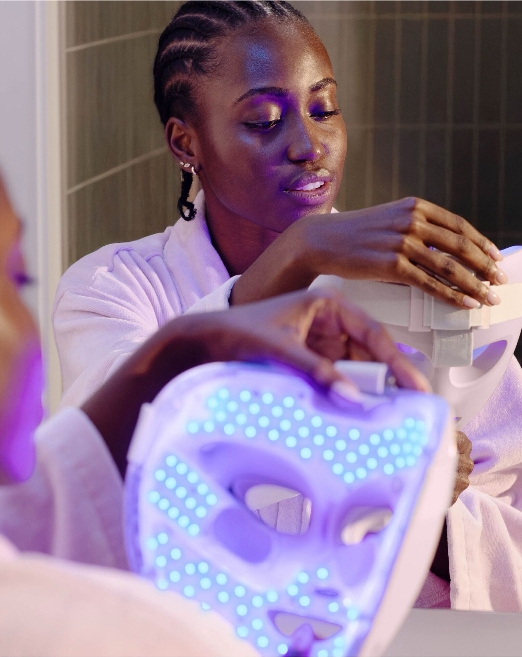 A woman in a white robe holding a glowing blue LED face mask in front of a mirror.