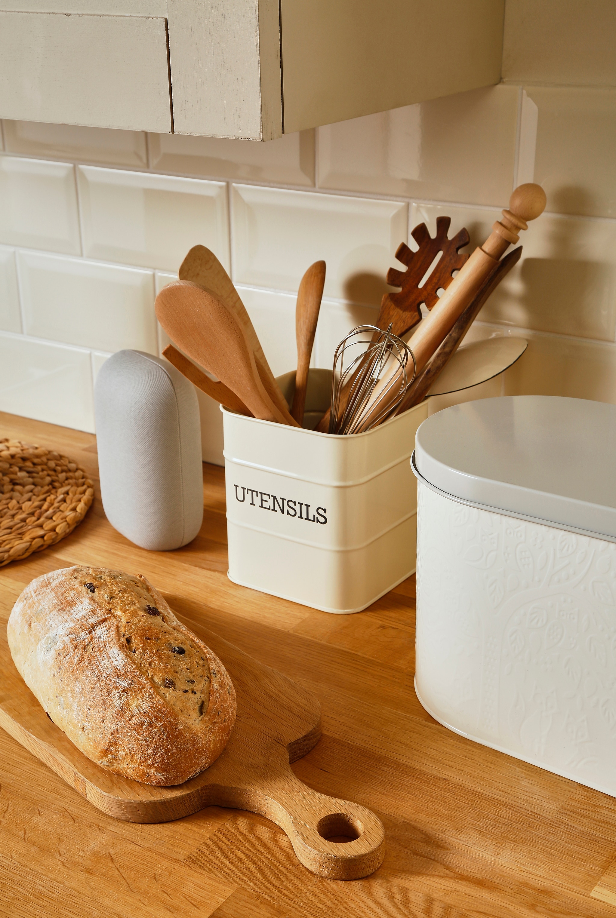 A kitchen worktop with some fresh bread on a chopping board. Utensils in a metal rectangular container displaying the word utensils on it written in captials.