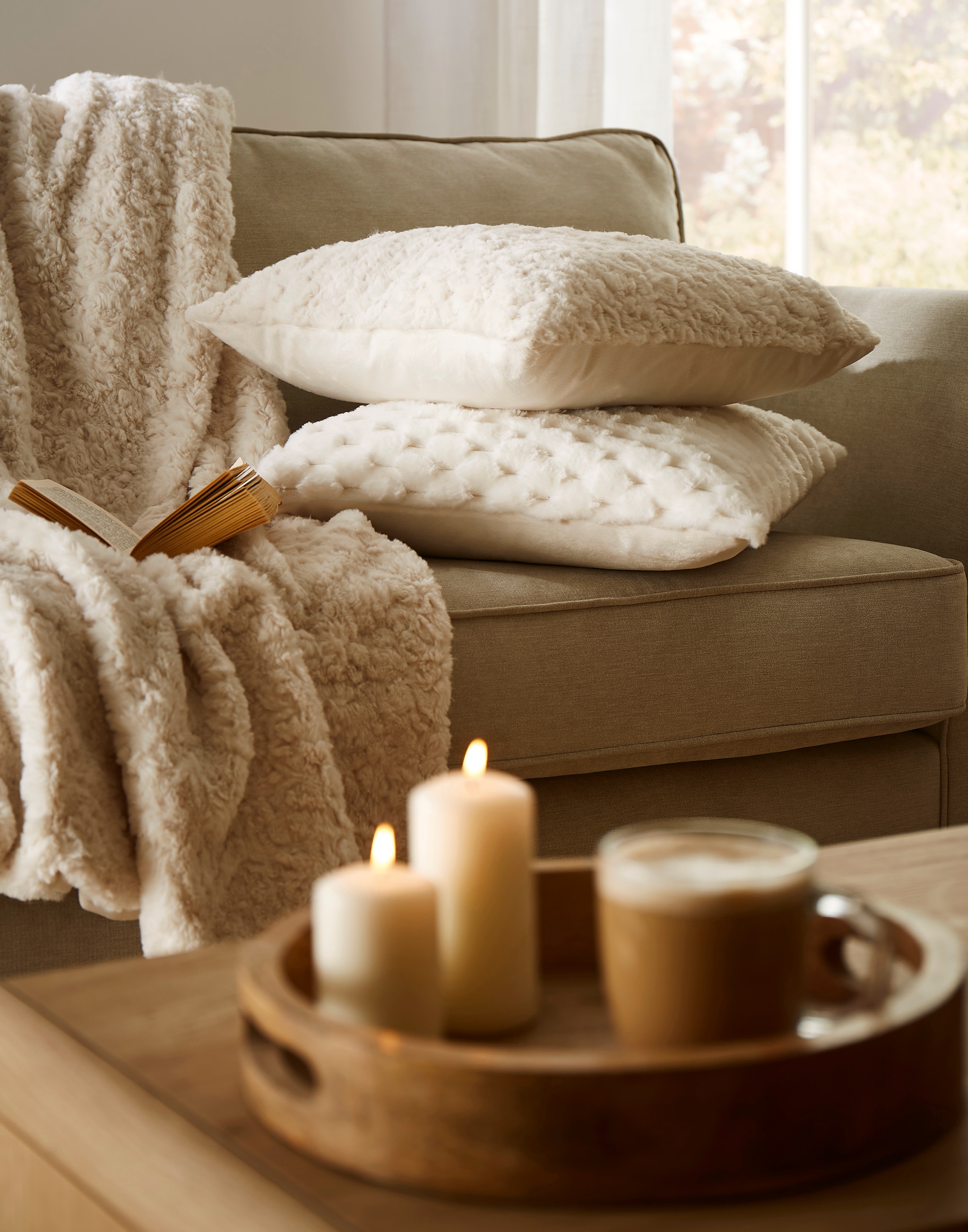 A cosy looking living rooms with cream and light brown throws and cushions. Candles lit on a table.