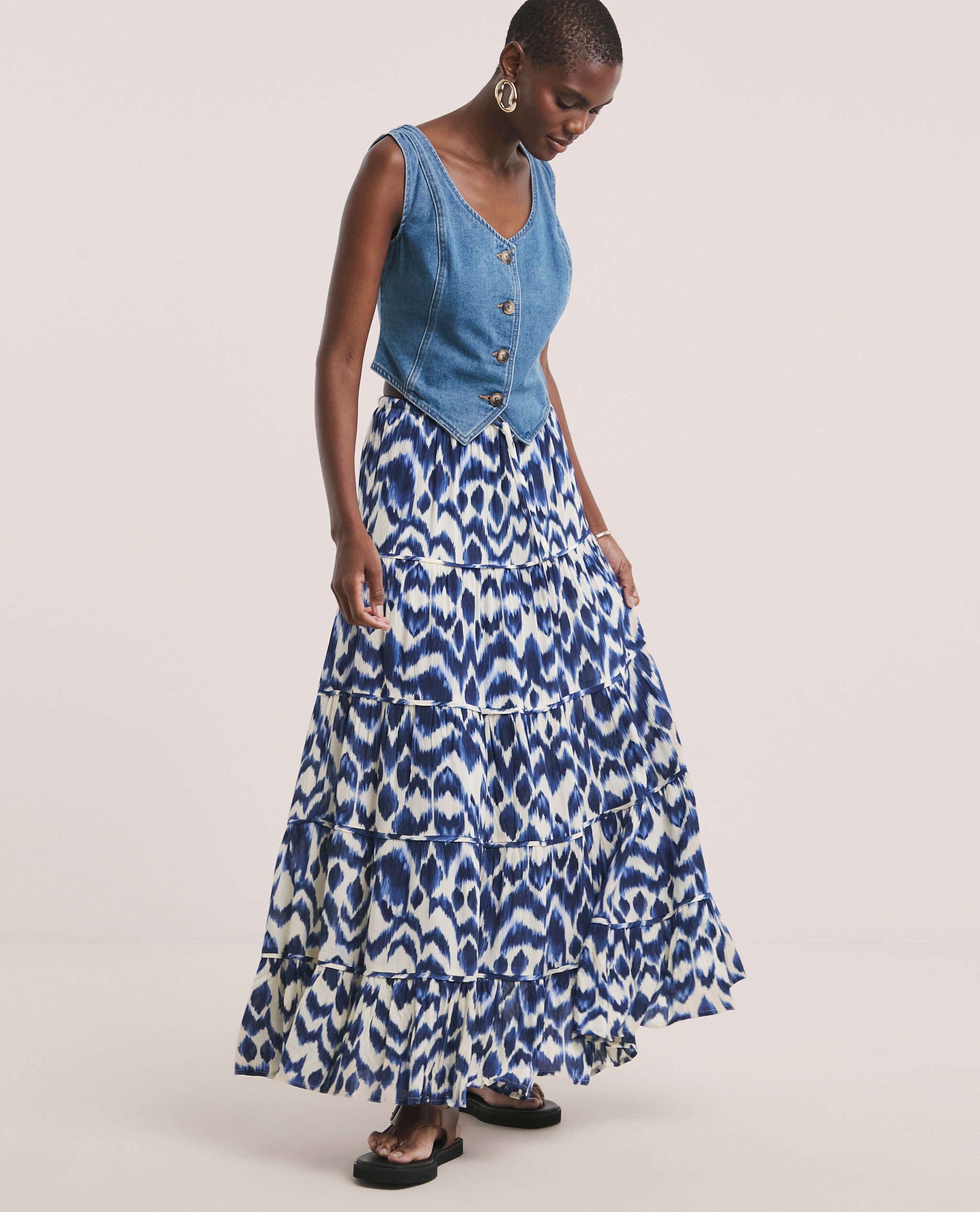 Model is wearing a blue denim waistcoat with a blue print textured crinkle tiered maxi skirt and chocolate toe post sandals.