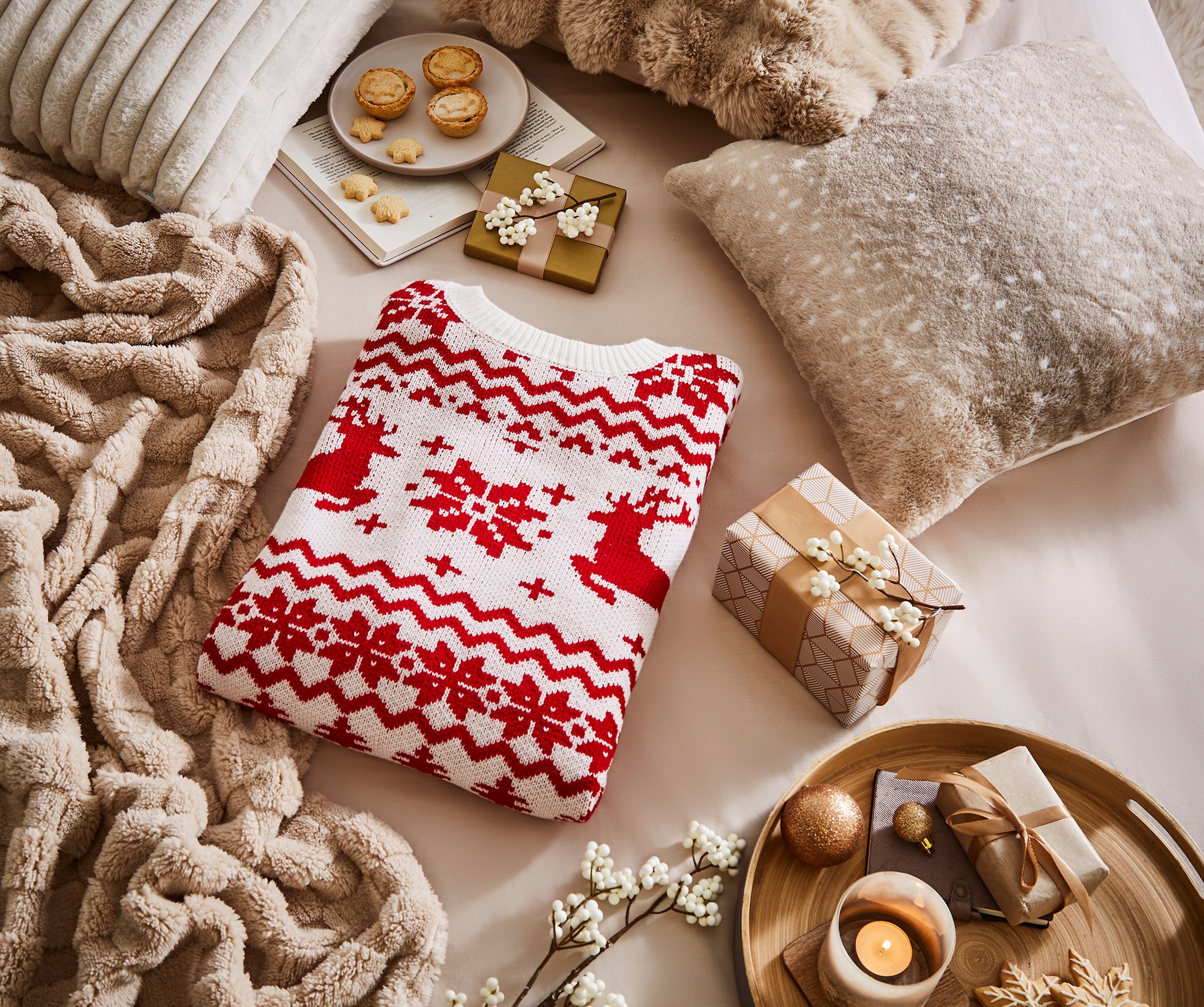 A folded FairIsle Reindeer Jumper on a table surrounded by a furry cushion, a faux fur throw and some seasonal Christmas food.