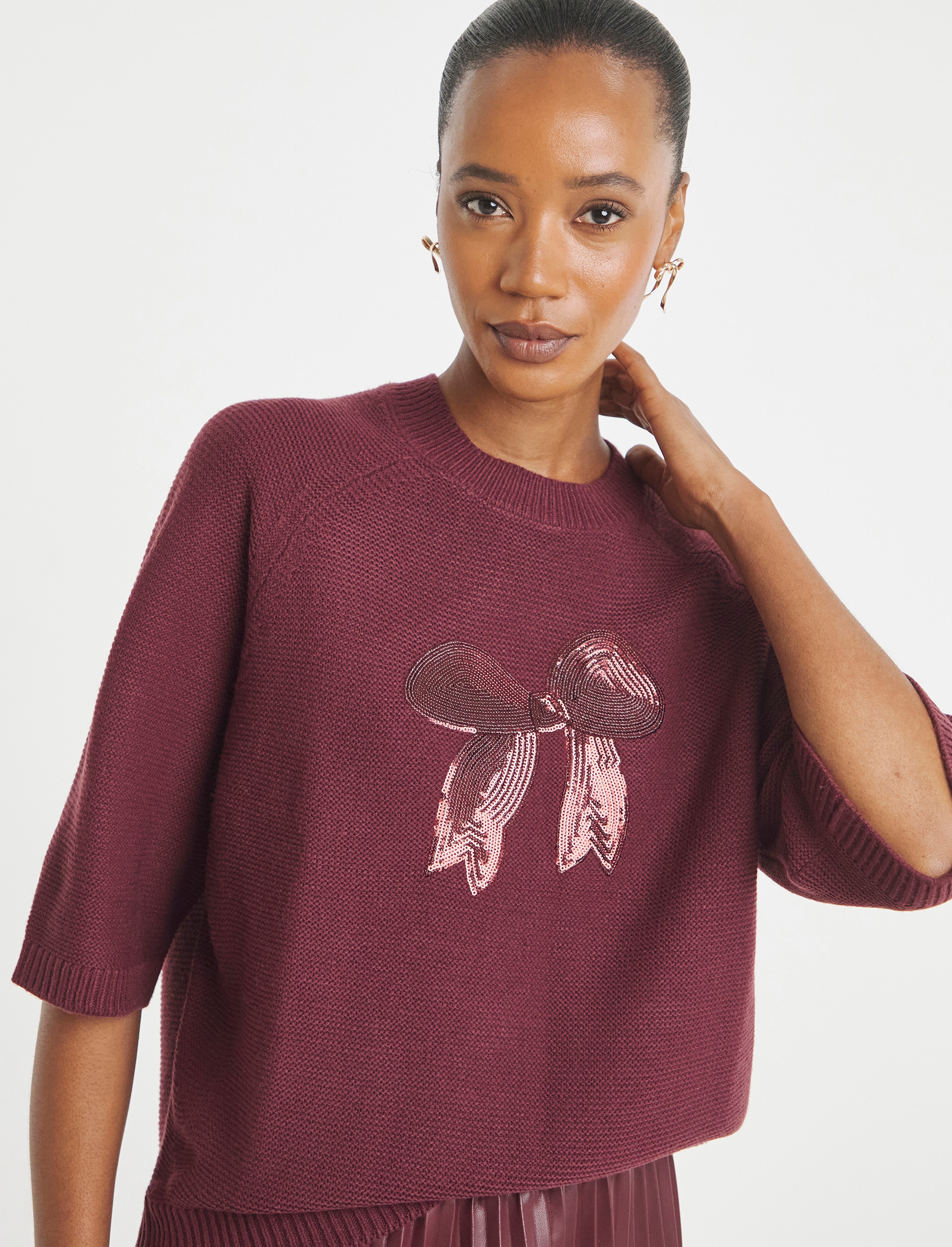A lady stood with her left arm touching her neck wearing a ruby coloured short sleeve jumper with a pink sequin embellished ribbon bow on the front