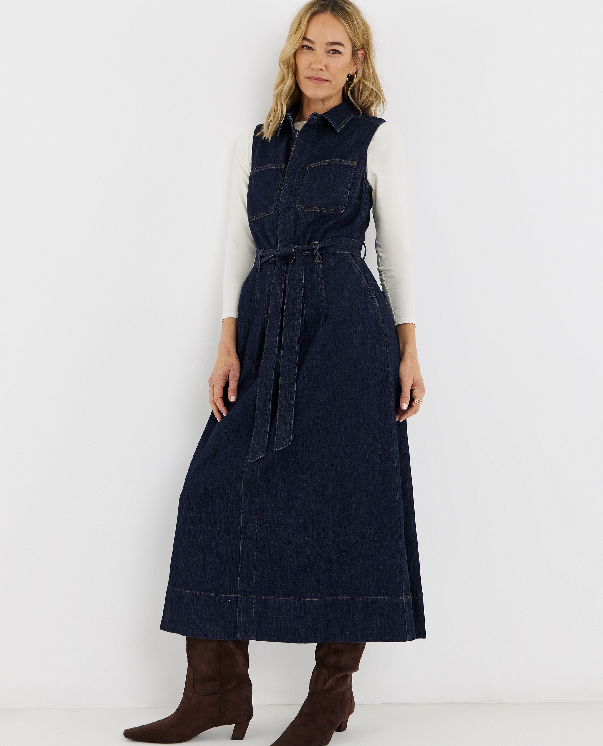 Model is wearing a white long sleeve top under a indigo sleeveless pocket front midaxi dress with a pair of chocolate boots.