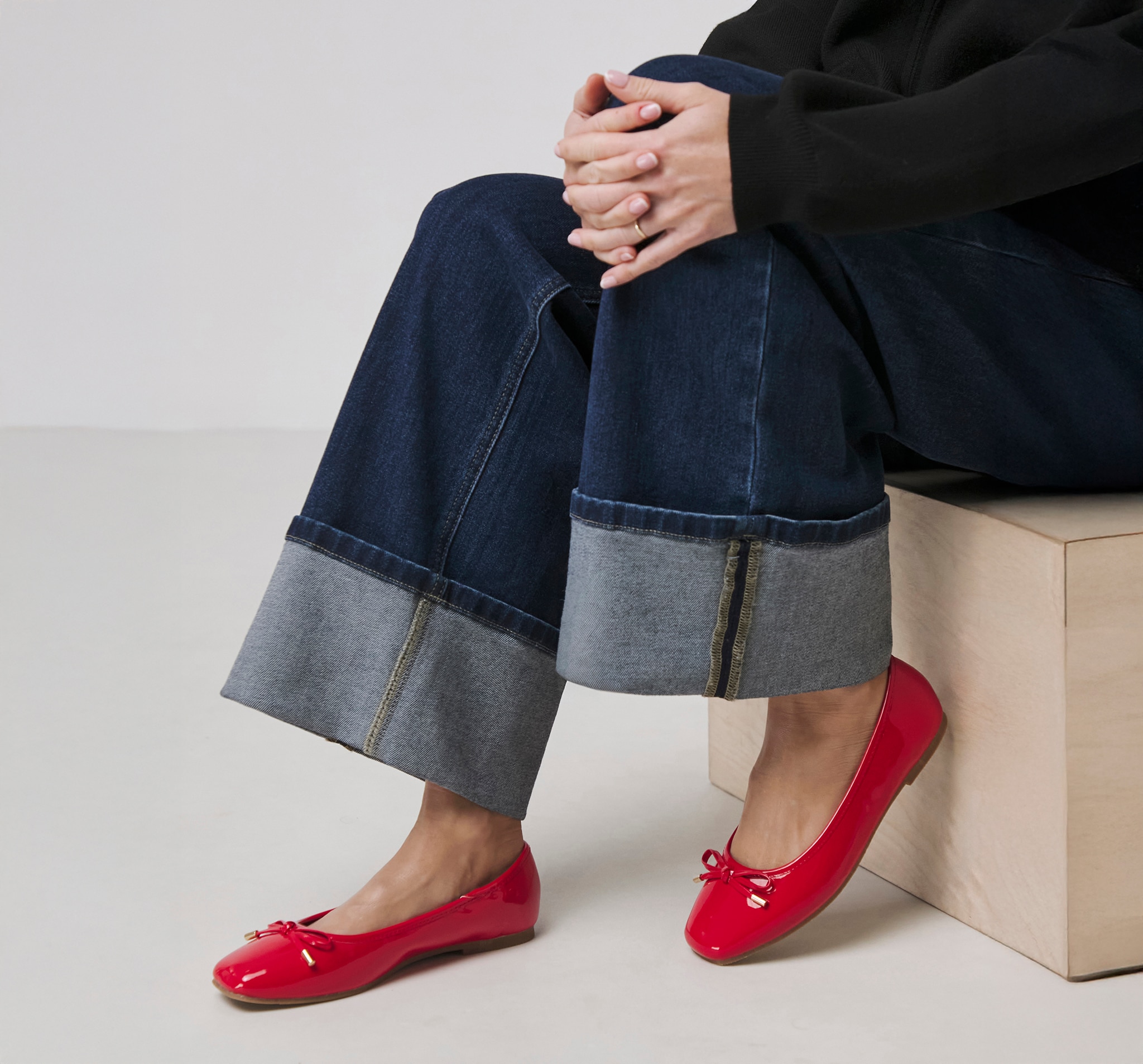 A person sat on a wooden box wearing indigo denim jeans with bright red patent Mary Jane shoes
