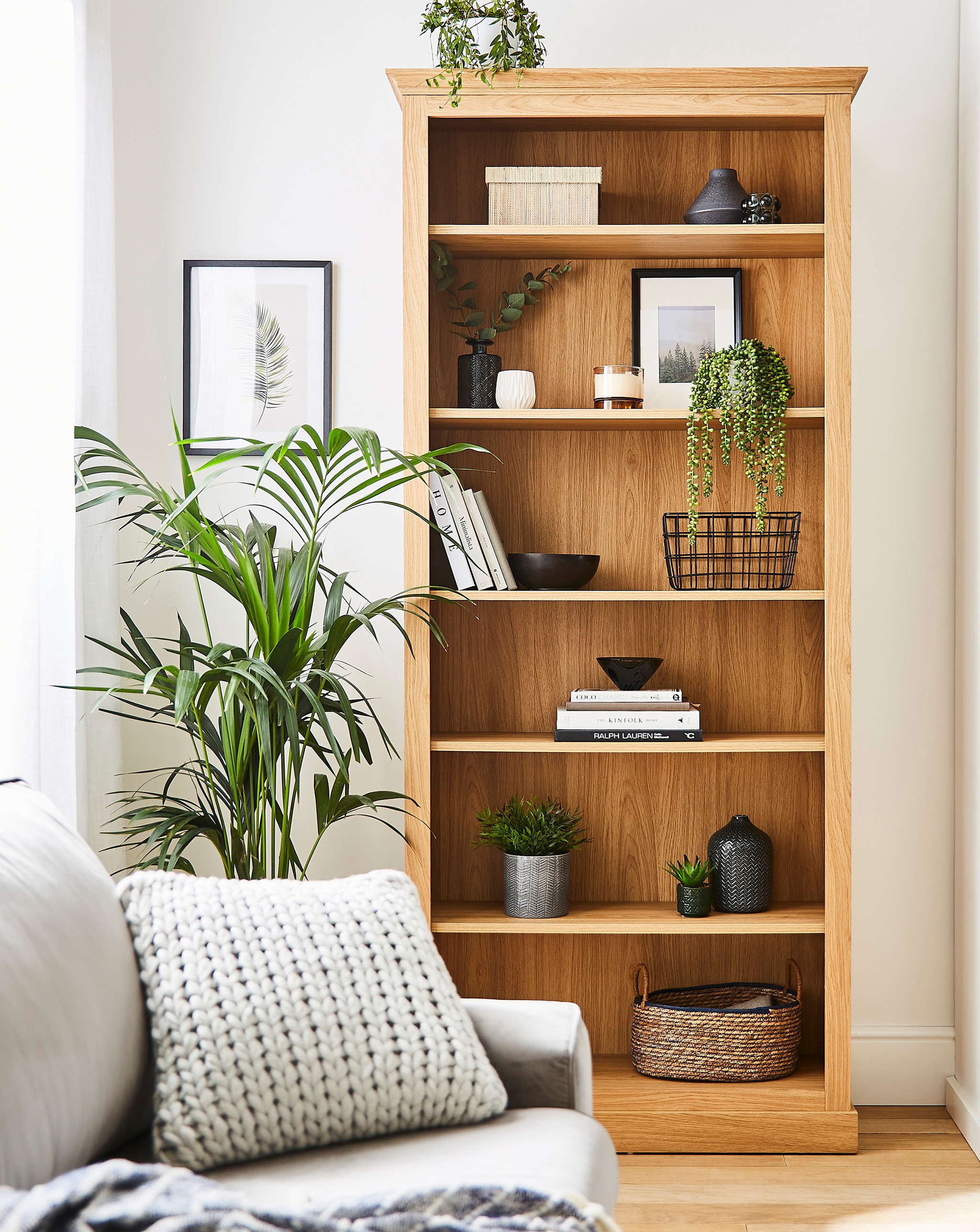 A modern living room with a tall wooden bookcase decorated with plants, books, and small ornaments beside a sofa with a chunky knitted cushion.