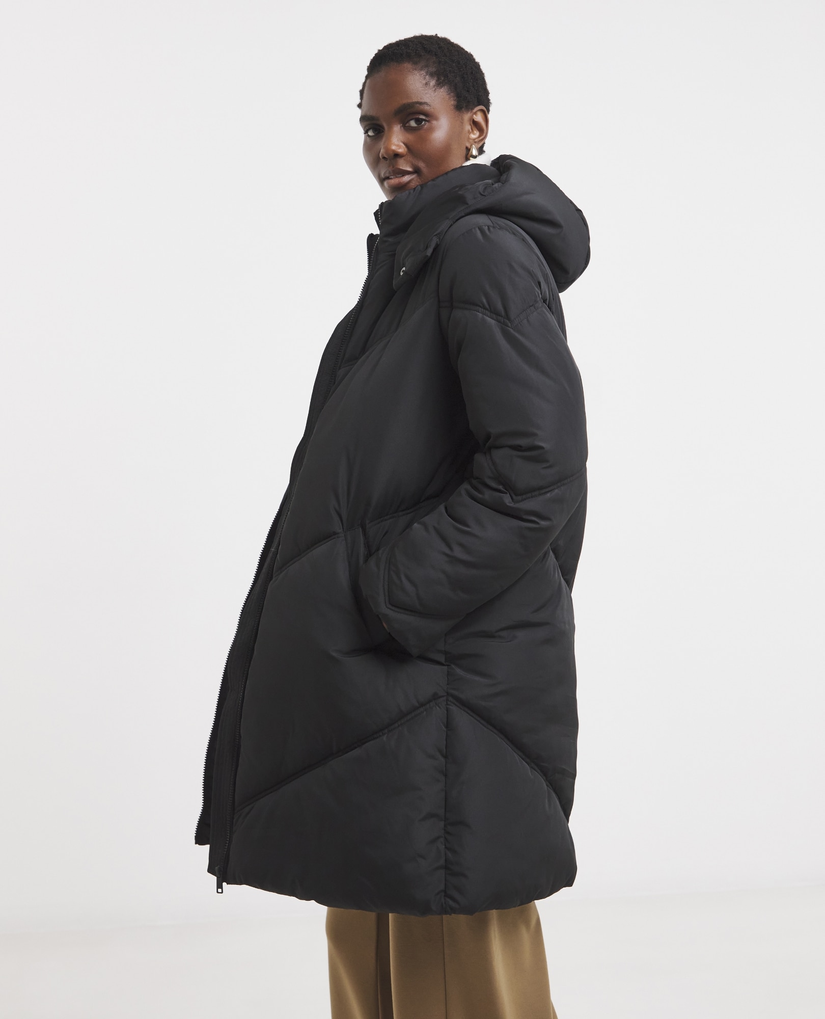 A woman with short hair wearing a long, black, oversized puffer coat with a hood and neutral-colored trousers